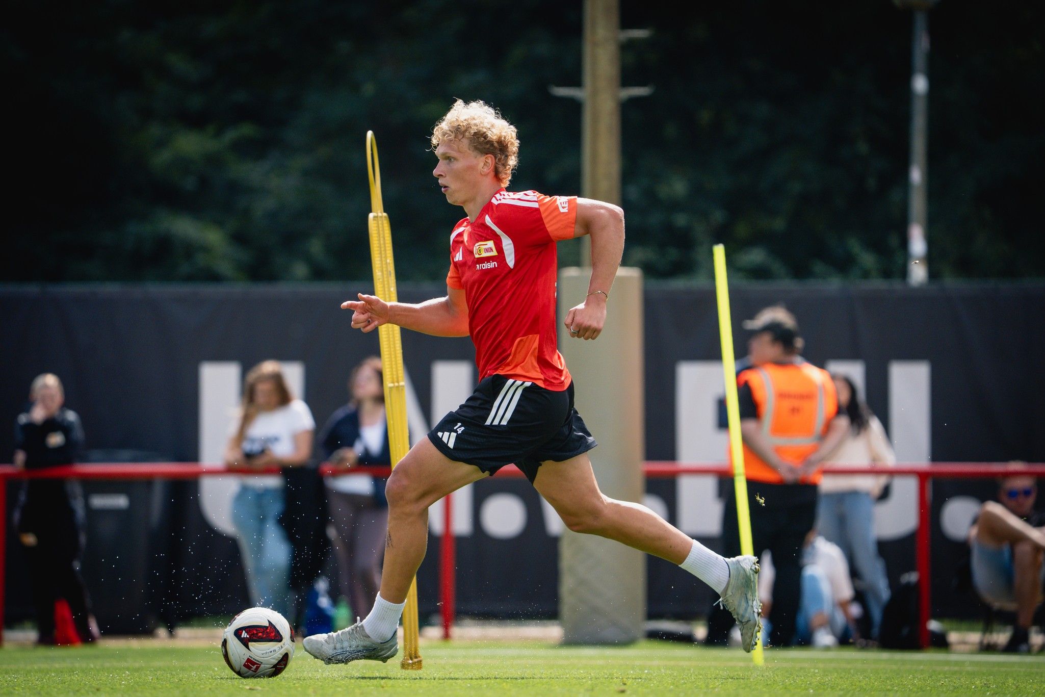 Ein junger Fußballspieler in rotem Trikot sprintet während des Trainings über das Spielfeld, umgeben von Pylonen.
