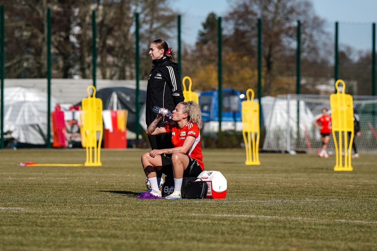Zwei Fußballspielerinnen auf dem Trainingsplatz; eine sitzt mit Wasserflasche, die andere steht daneben. Gelbe Trainingshilfen im Hintergrund.