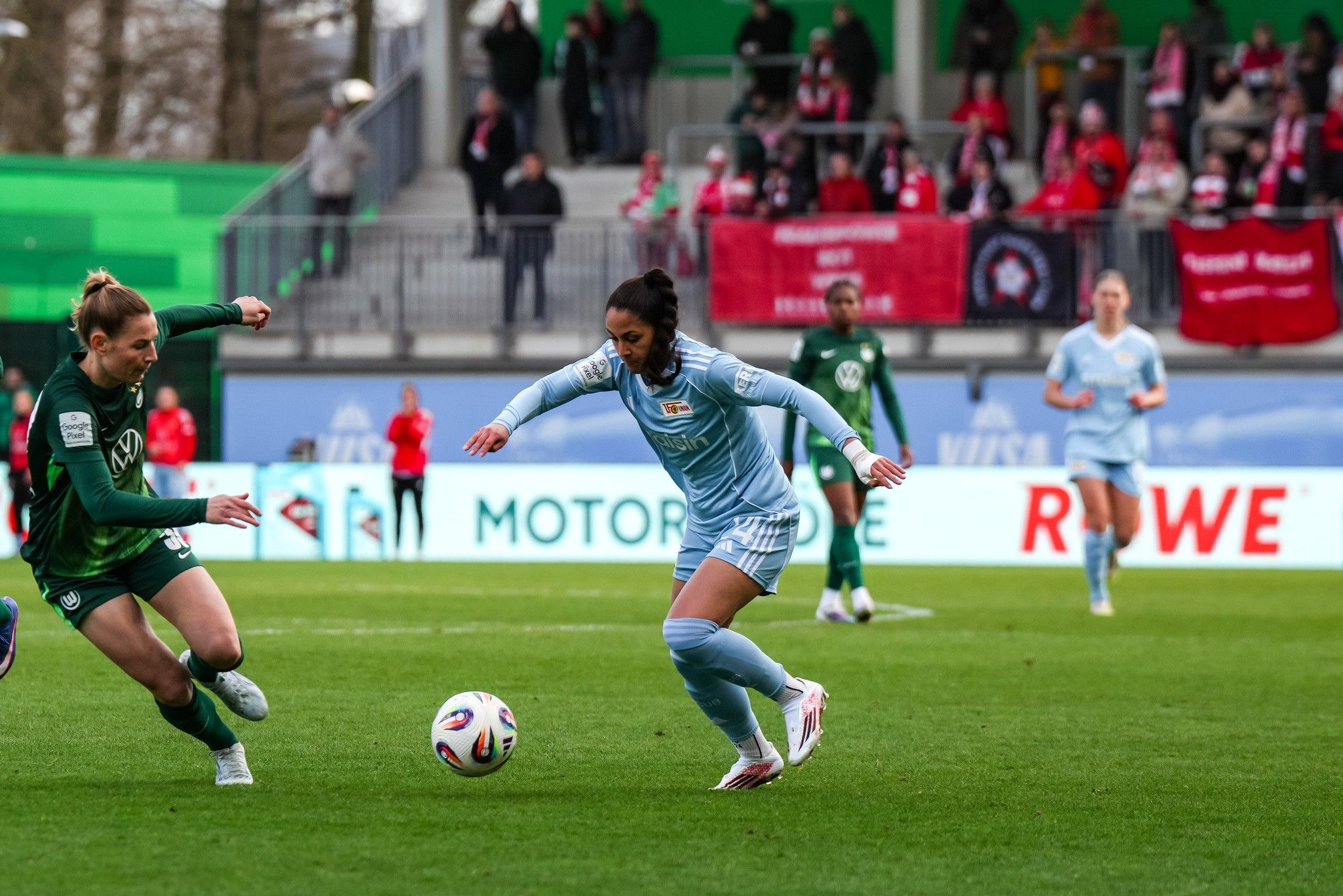 Eine Spielerin in hellblau dribbelt den Ball, während eine gegnerische Spielerin in grün versucht, ihn ihr abzunehmen.