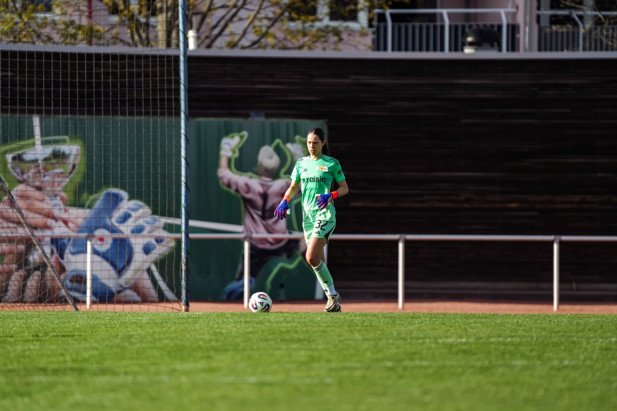 Eine Fußballtorhüterin in grüner Ausrüstung steht auf dem Spielfeld, im Hintergrund ist ein muralartiges Sportmotiv.