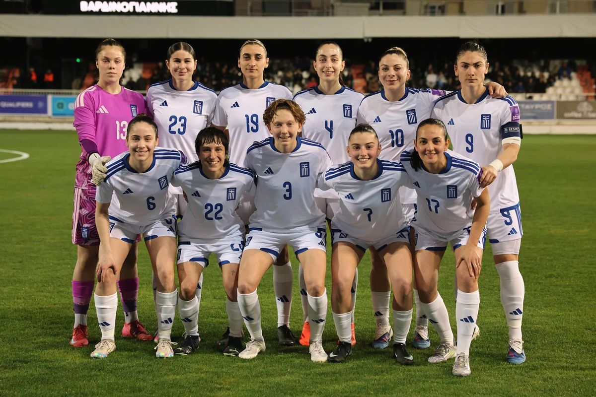 Griechische Frauenfußballmannschaft posiert in weißen Trikots auf einem Fußballplatz vor einer Tribüne.