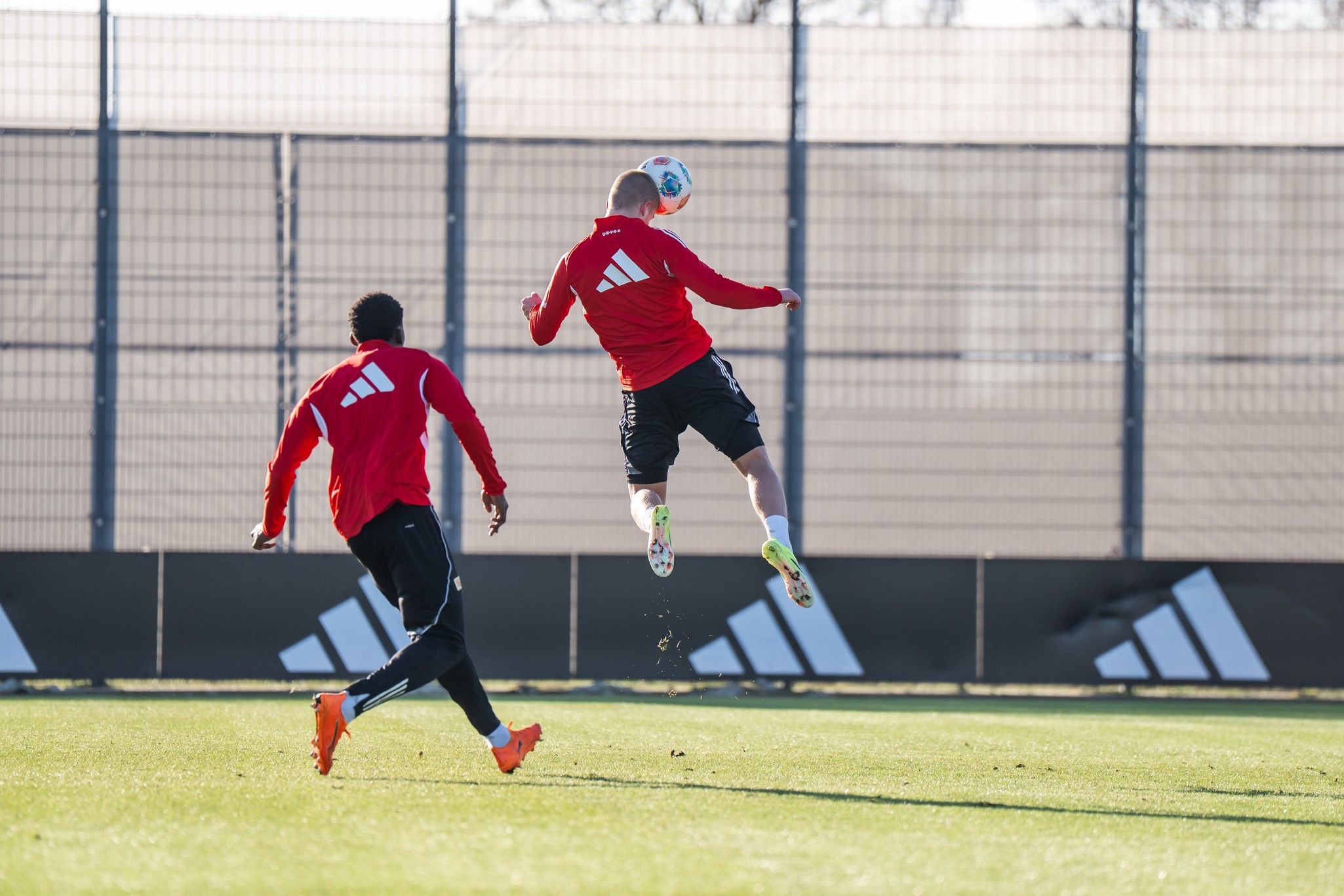 Zwei Fußballspieler in roten Trikots üben auf einem Trainingsplatz, während einer den Ball mit einem Sprung spielt.