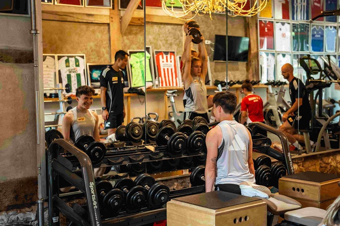 Mehrere Personen im Fitnessstudio heben Gewichte und trainieren vor einem Spiegel, umgeben von Fitnessgeräten und Sporttrikots.