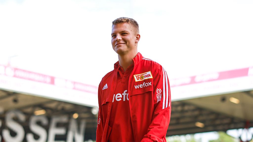 Ein junger Mann in roter Sportjacke lächelt auf einem Fußballplatz, im Hintergrund sind Tribünen zu sehen.