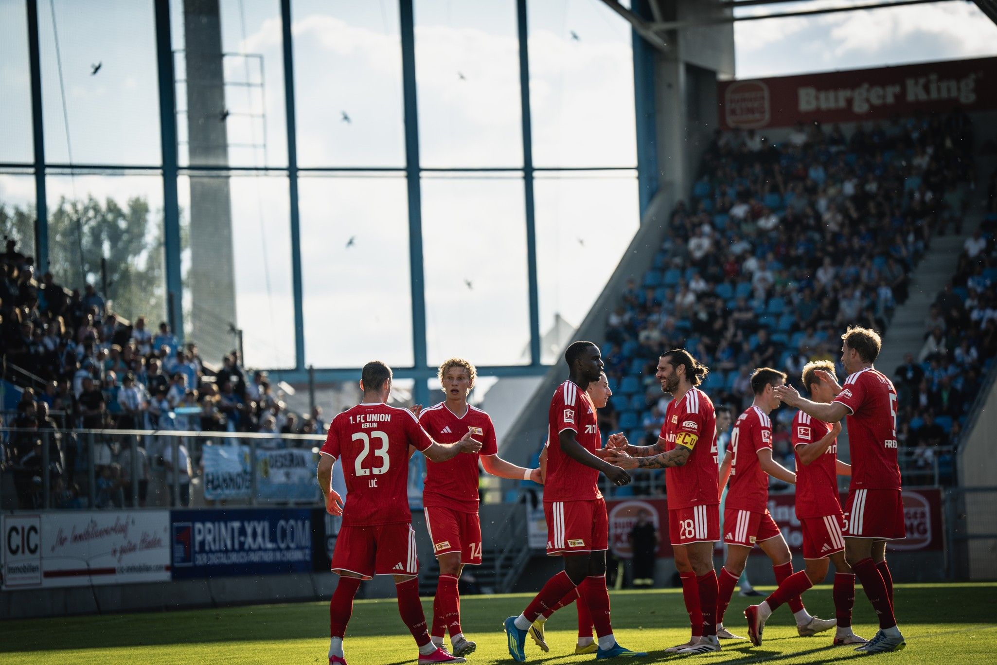Spieler in roten Trikots feiern ein Tor in einem Stadion mit Zuschauern im Hintergrund.
