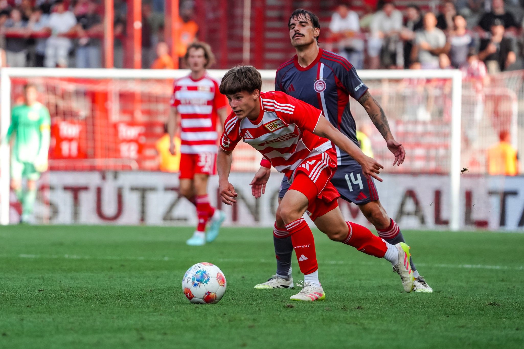 Ein Fußballspieler in roten Trikots verfolgt den Ball, während ein Spieler in grauem Trikot ihn bedrängt. Zuschauer im Hintergrund.
