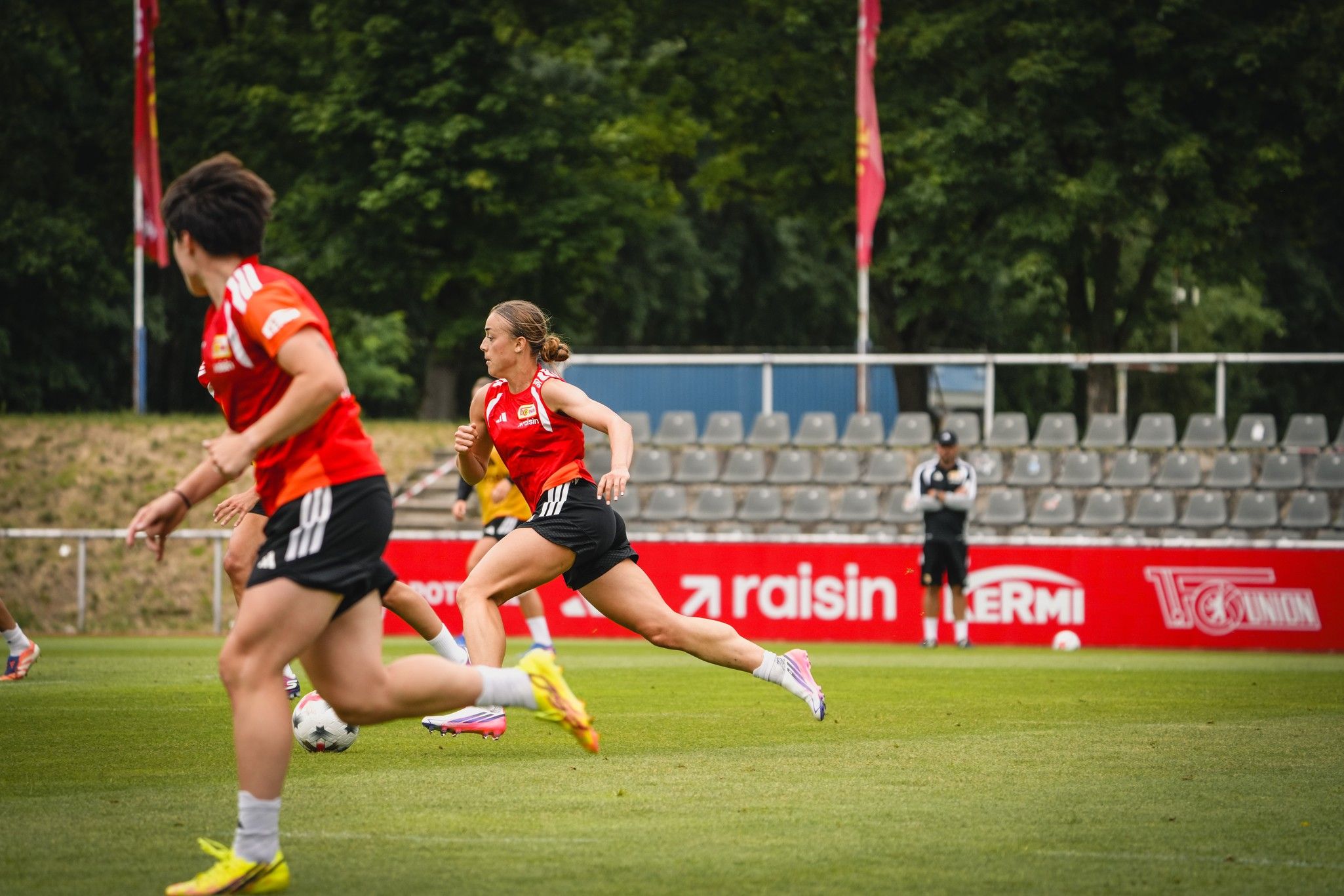 Zwei Fußballspielerinnen in roten Trikots trainieren auf einem grünen Platz mit Tribünen im Hintergrund.