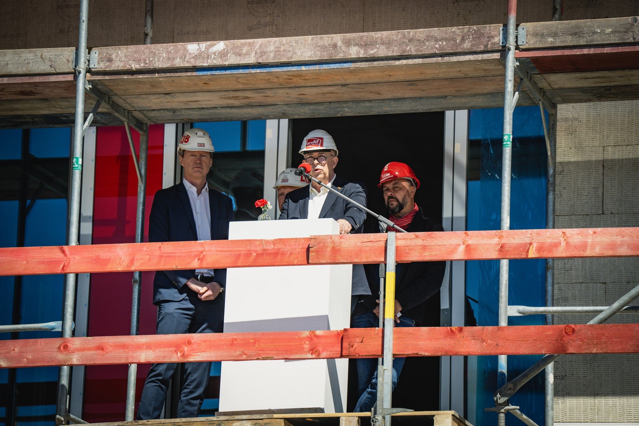Männer in Sicherheitshelmen stehen auf einem Gerüst und sprechen vor einer weißen Säule, umgeben von Baustellenmaterial.