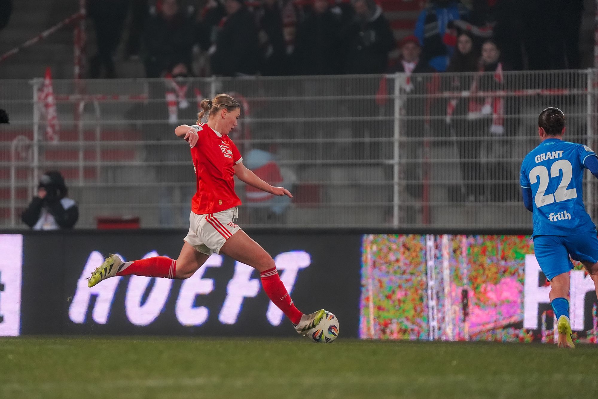 Spielszene aus einem Fußballspiel, eine Spielerin in rotem Trikot tritt den Ball, ein weiterer Spieler in blauem Trikot steht bereit.