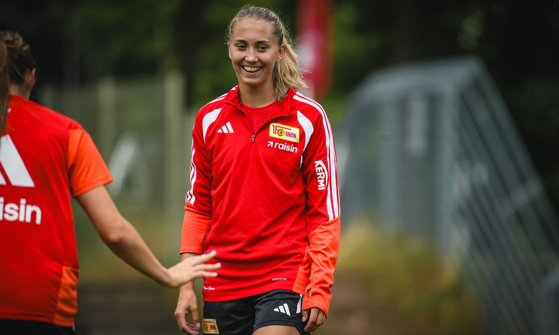 Eine lächelnde Sportlerin in einem roten Trikot bei einem Training, umgeben von anderen Spielerinnen.