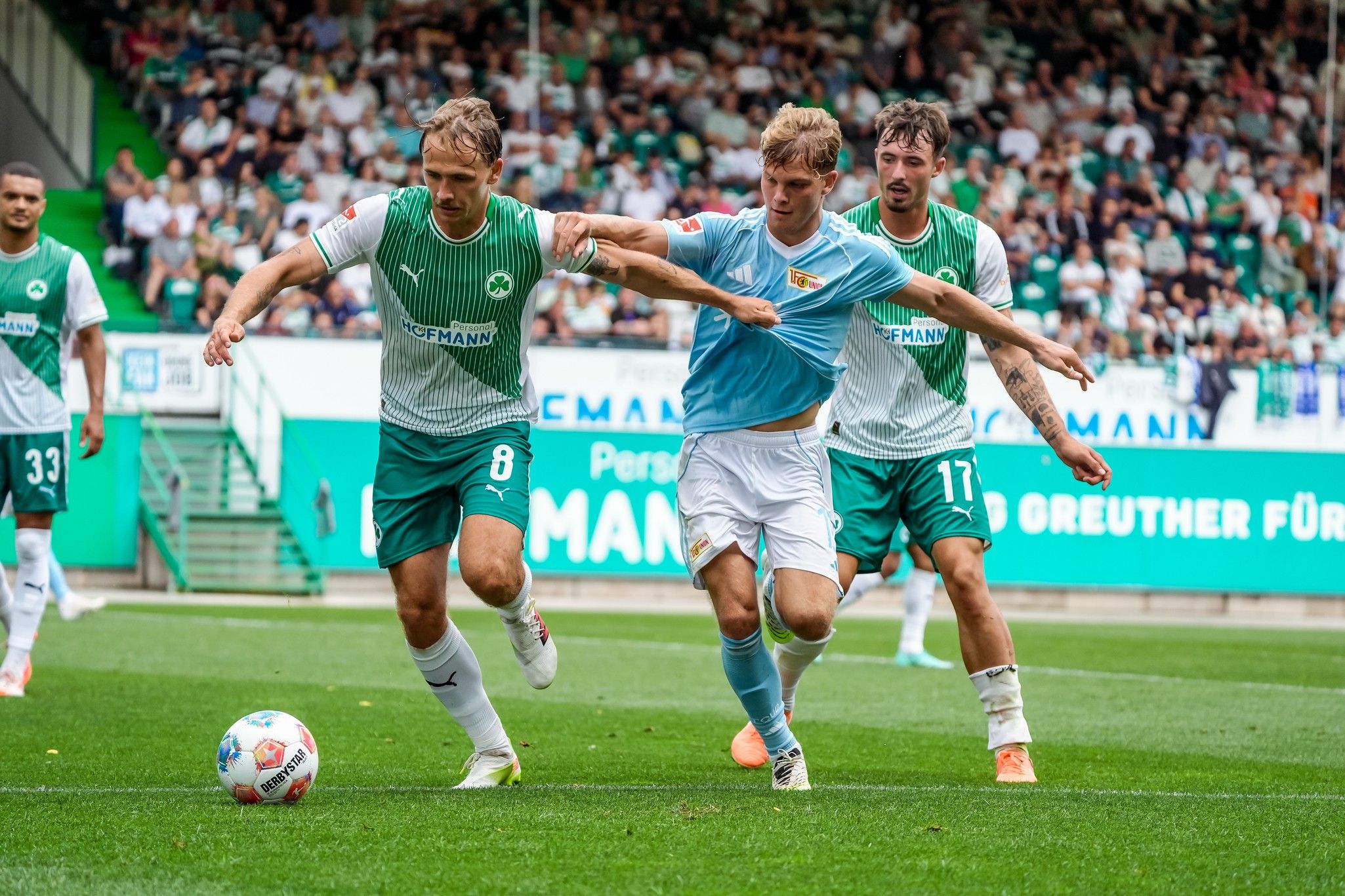 Spielszene im Fußball, zwei Spieler kämpfen um den Ball, Zuschauer im Hintergrund, Stadionatmosphäre.