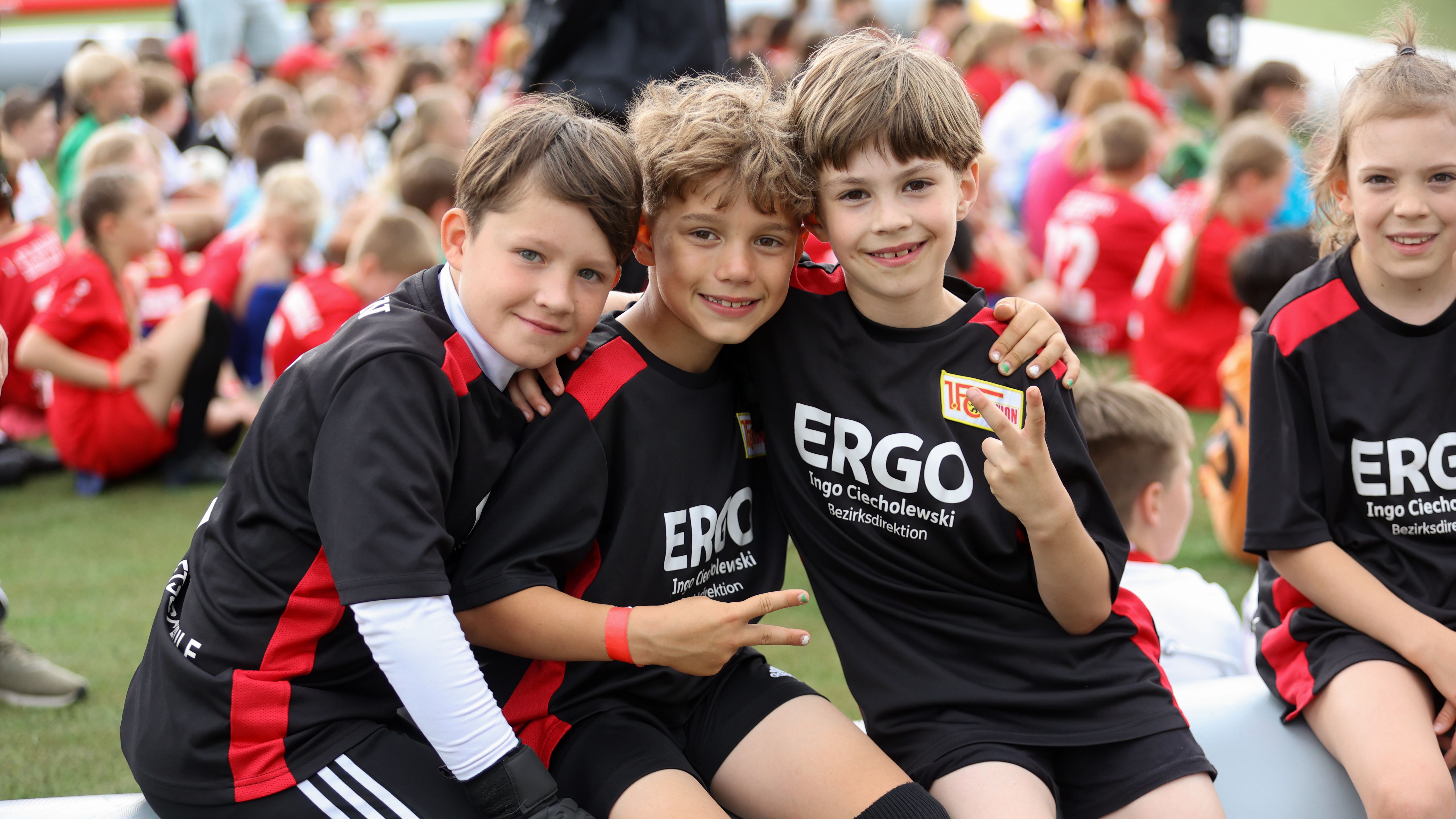 Drei Kinder in Fußballtrikots sitzen auf einer Bank, umgeben von anderen Kindern auf einem Sportplatz.