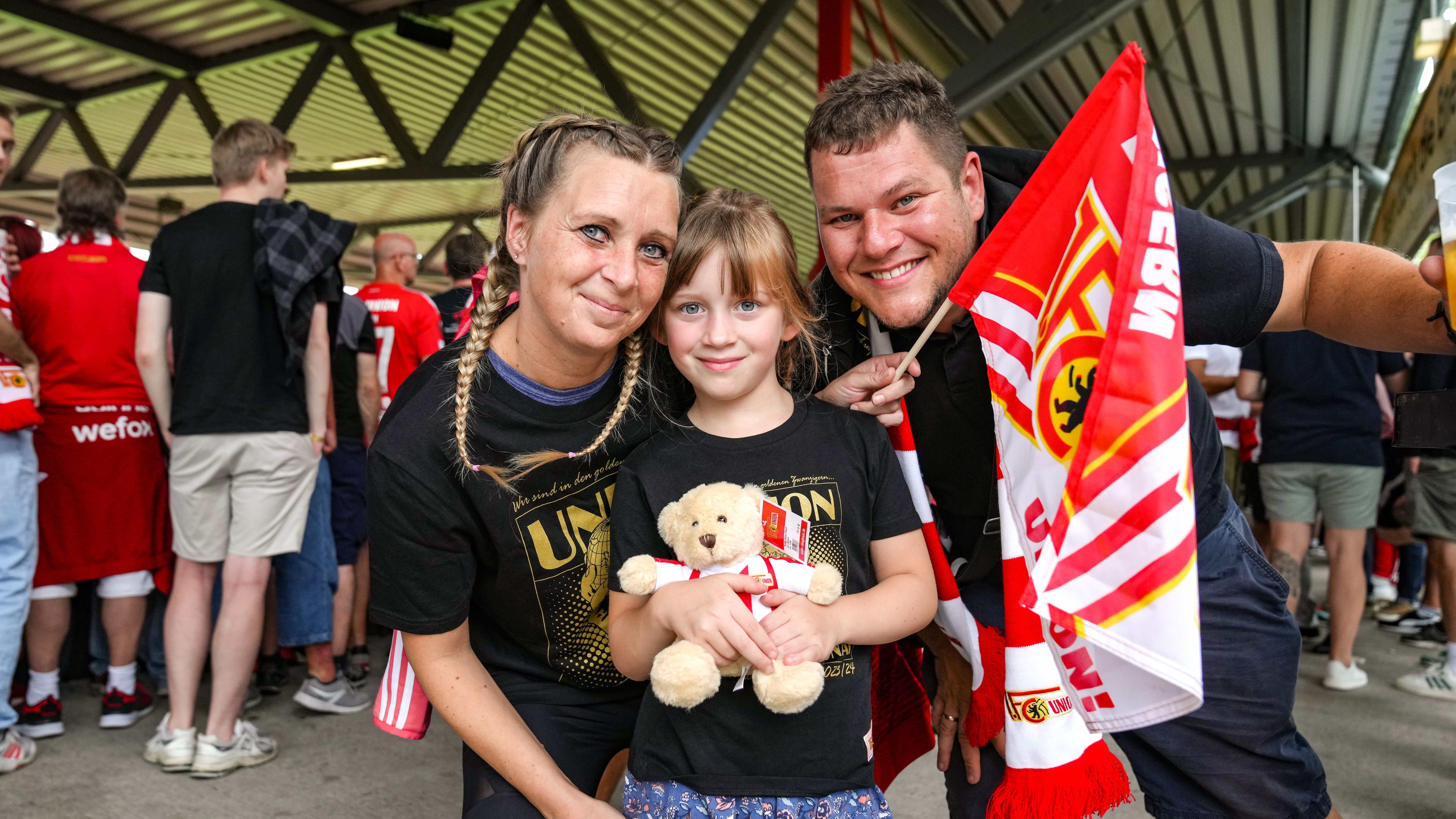 Eine Frau und ein Mann posieren mit einem Mädchen, das ein Stofftier hält, umgeben von Fans in rot-weißen Trikots.