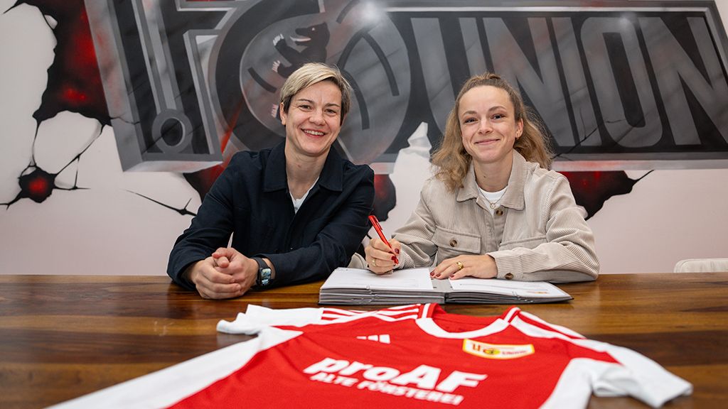 Two women are sitting at a table signing a contract. In the foreground, there is a red soccer jersey.