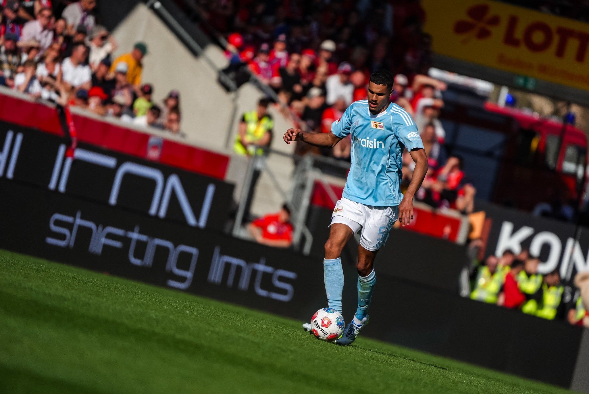 Spieler in hellblauem Trikot dribbelt den Ball auf dem Spielfeld, umgeben von Fans und Sicherheitskräften.