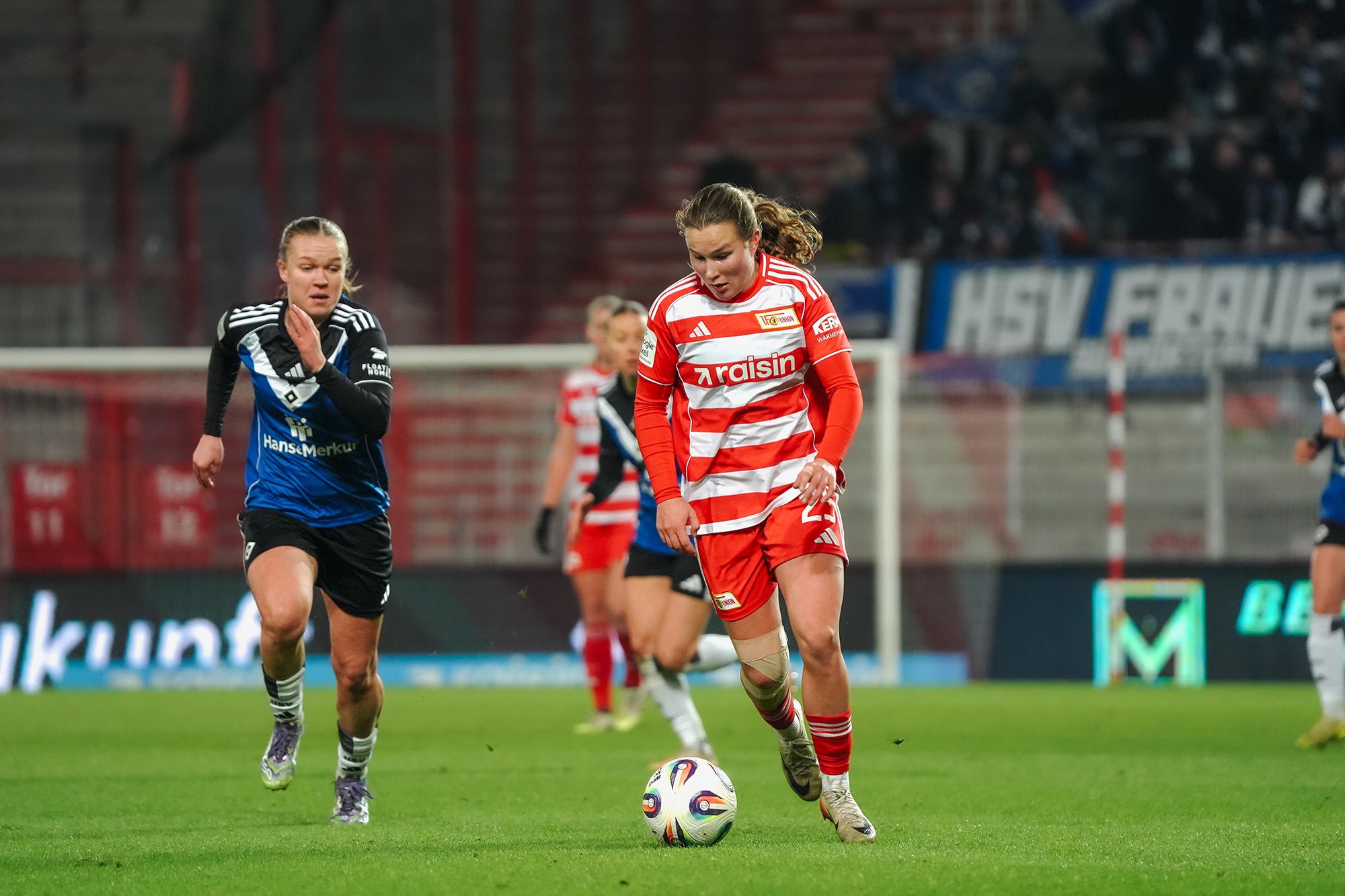 Spielszene im Frauenfußball: Eine Spielerin in rot-weißer Uniform dribbelt den Ball, während eine Gegnerin in blauer Uniform sie verfolgt.