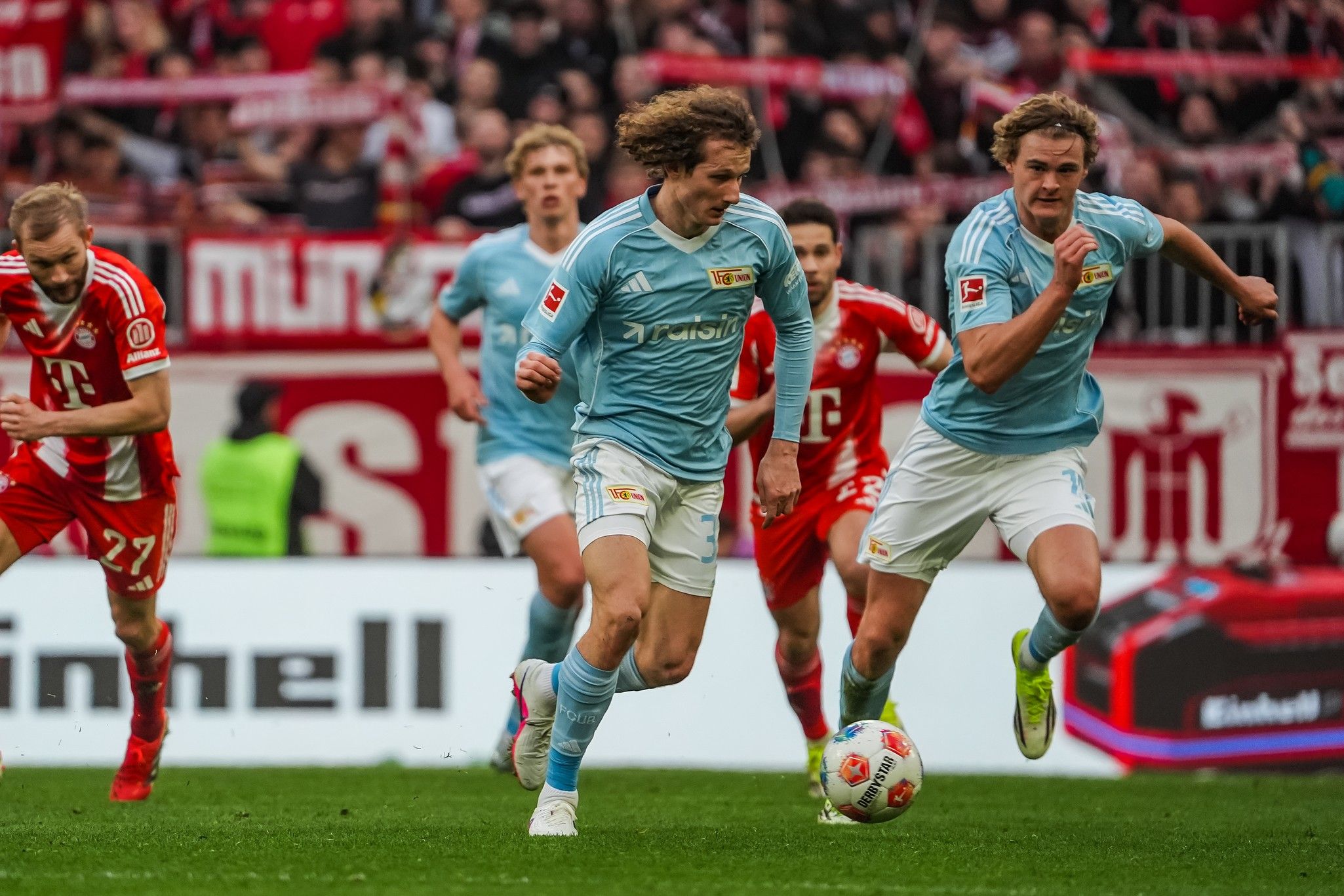 Spielszene aus einem Fußballspiel: Zwei Spieler in blauen Trikots sprinten mit dem Ball, während ein Gegner in rotem Trikot verfolgt.