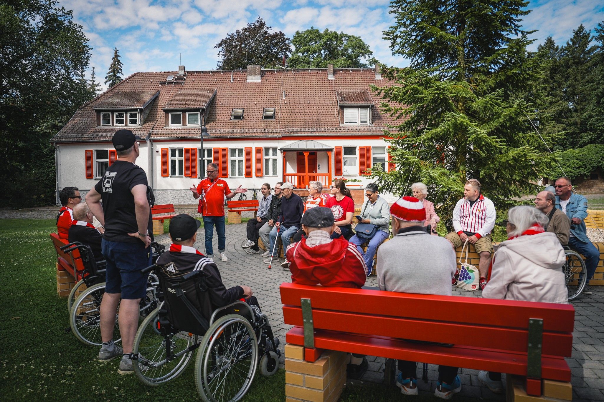 Eine Gruppe von Menschen sitzt im Freien auf Bänken, während eine Person in roter Kleidung spricht. Im Hintergrund ein Gebäude mit roten Fensterläden.