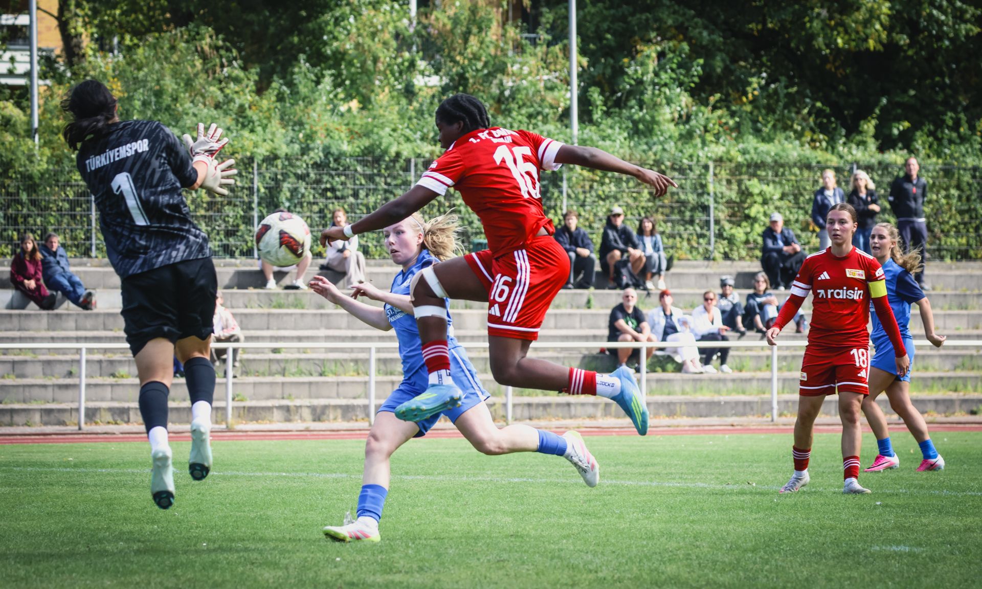 Eine Spielerin in rotem Trikot springt hoch, um den Ball in einem Fußballspiel zu erreichen, während die Torhüterin und andere Spielerinnen zuschauen.