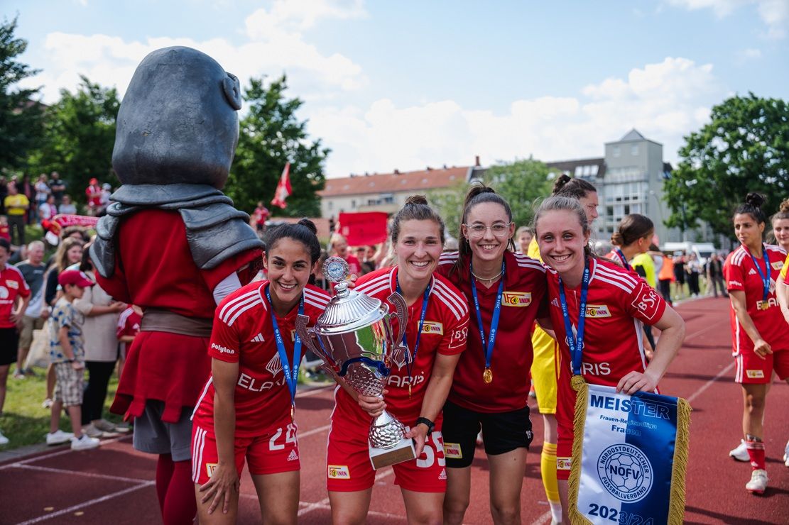 Vier Fußballspielerinnen in roten Trikots halten einen Meisterschaftspokal und eine Medaille, umgeben von jubelnden Fans.