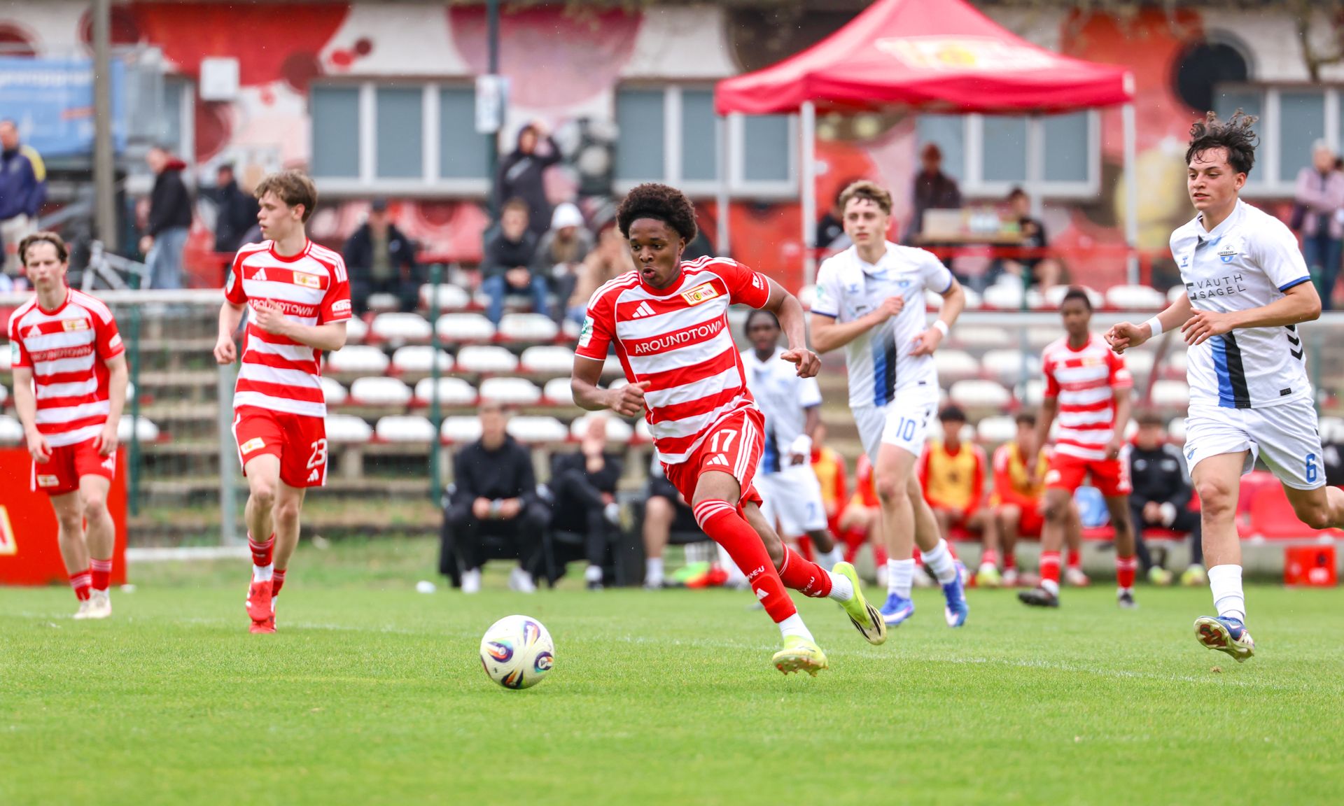 Ein Fußballspieler mit roten und weißen Streifen läuft dribbelnd über ein Spielfeld, während andere Spieler und Zuschauer im Hintergrund zu sehen sind.