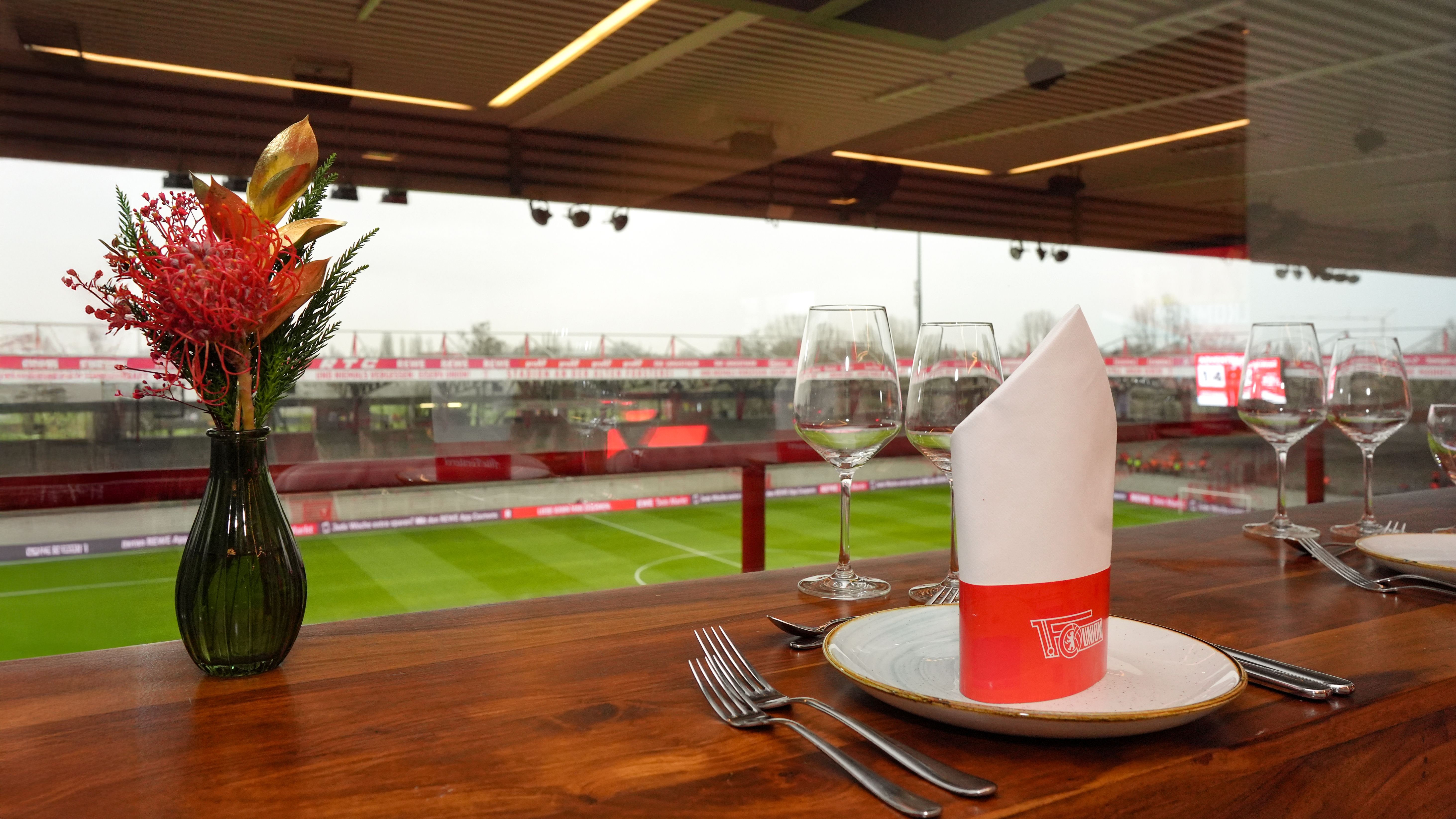 Ein gedeckter Tisch mit einem Teller, Besteck und Glas in einem Restaurant mit Blick auf ein Fußballfeld.