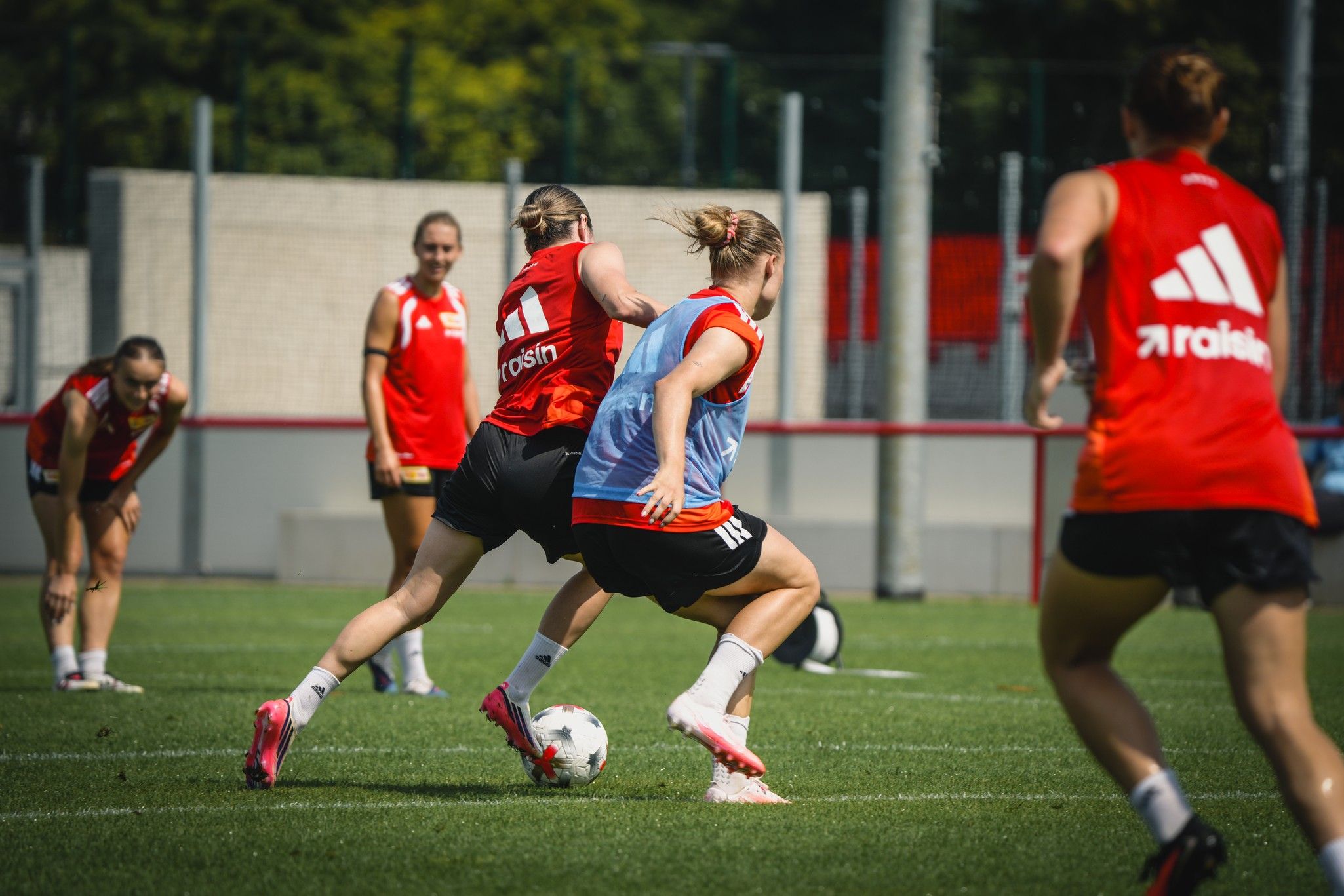Zwei Fußballspielerinnen im Training dribbeln den Ball auf einem Rasenplatz, während andere im Hintergrund Übungen machen.