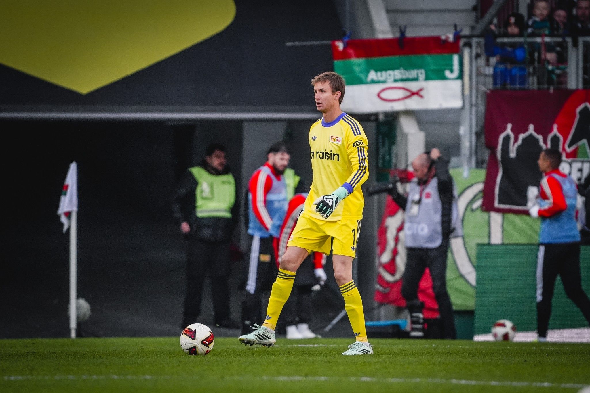 Torwart in gelber Ausrüstung steht im Stadion, im Hintergrund sind Fans und Fahnen zu sehen.