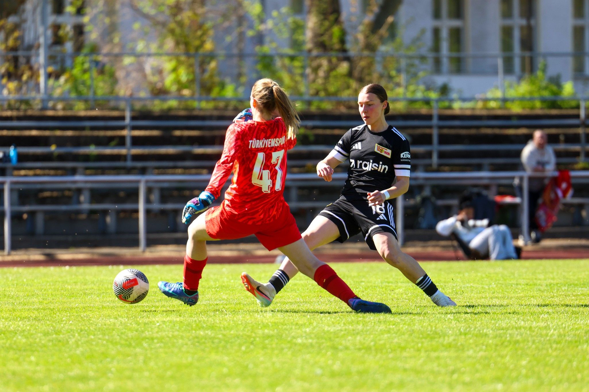 Eine Torwartin in roter Kleidung versucht, den Ball zu halten, während eine Spielerin in schwarzer Kleidung angreift.