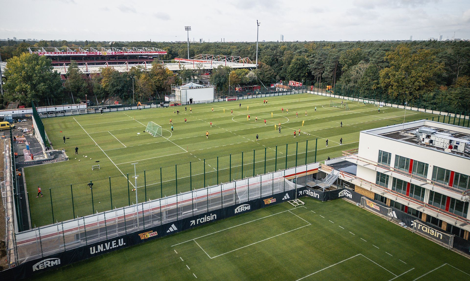 Ein Fußballfeld mit Spielern in Aktion. Im Hintergrund sind Übungseinrichtungen und ein Stadion zu sehen.