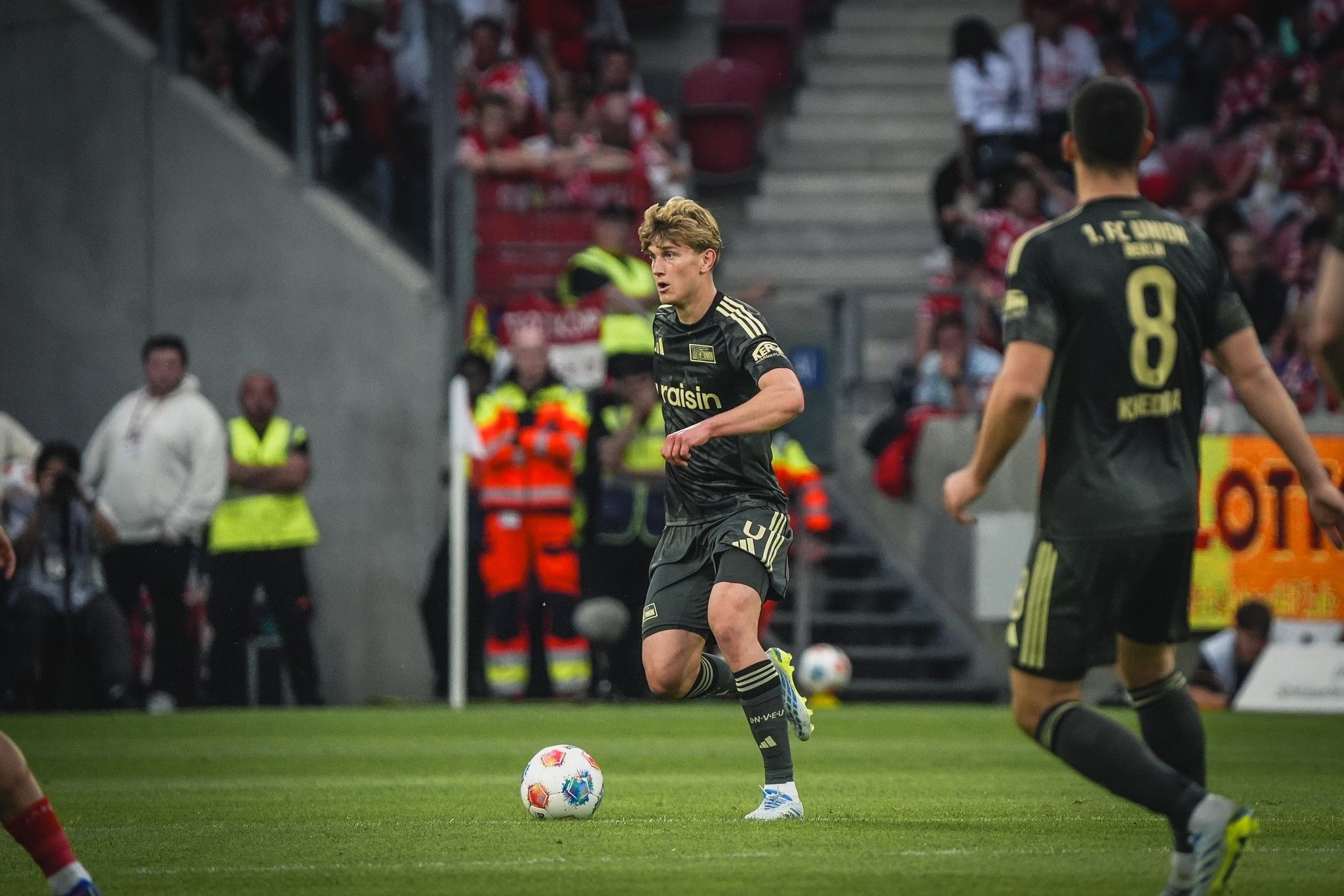 Ein Fußballspieler in grauer Uniform dribbelt den Ball, während ein zweiter Spieler in der Nähe steht. Menschenmengen im Hintergrund.