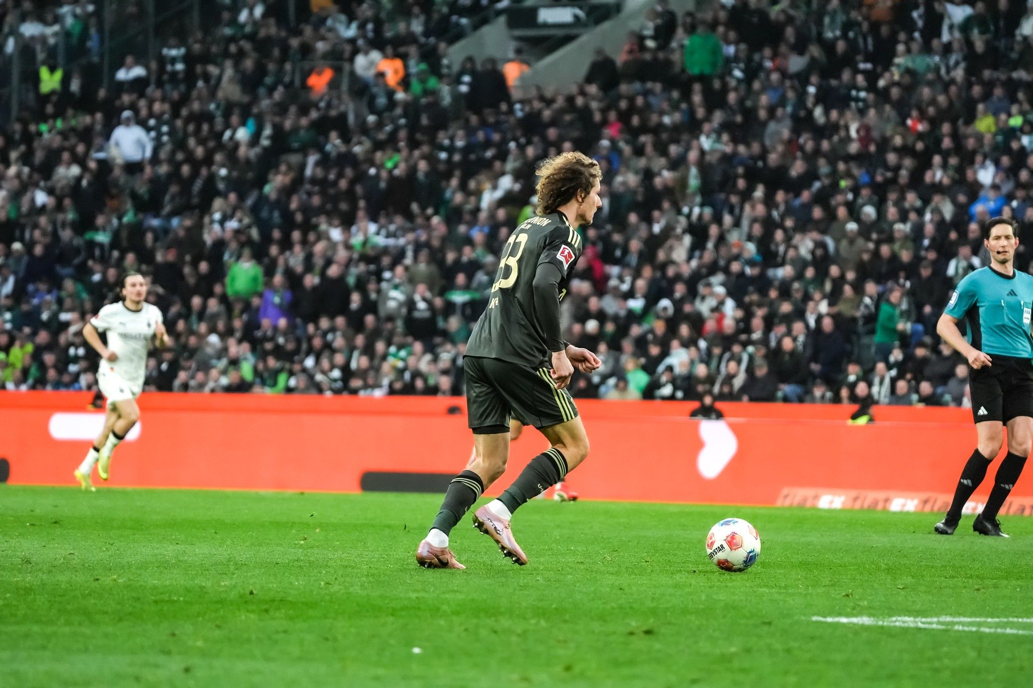 Ein Fußballspieler mit der Nummer 13 dribbelt den Ball auf einem Sportfeld, während Zuschauer im Hintergrund jubeln.