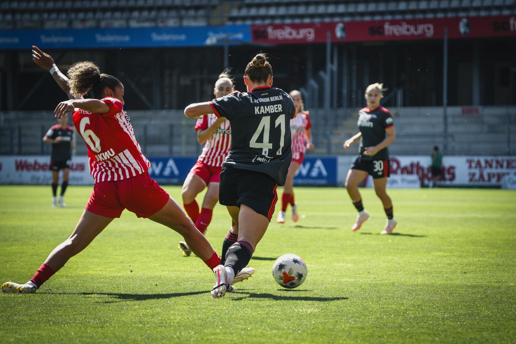 Spielszene im Fußball mit Spielerinnen in roten und schwarzen Trikots; eine Spielerin dribbelt den Ball, während eine andere attackiert.