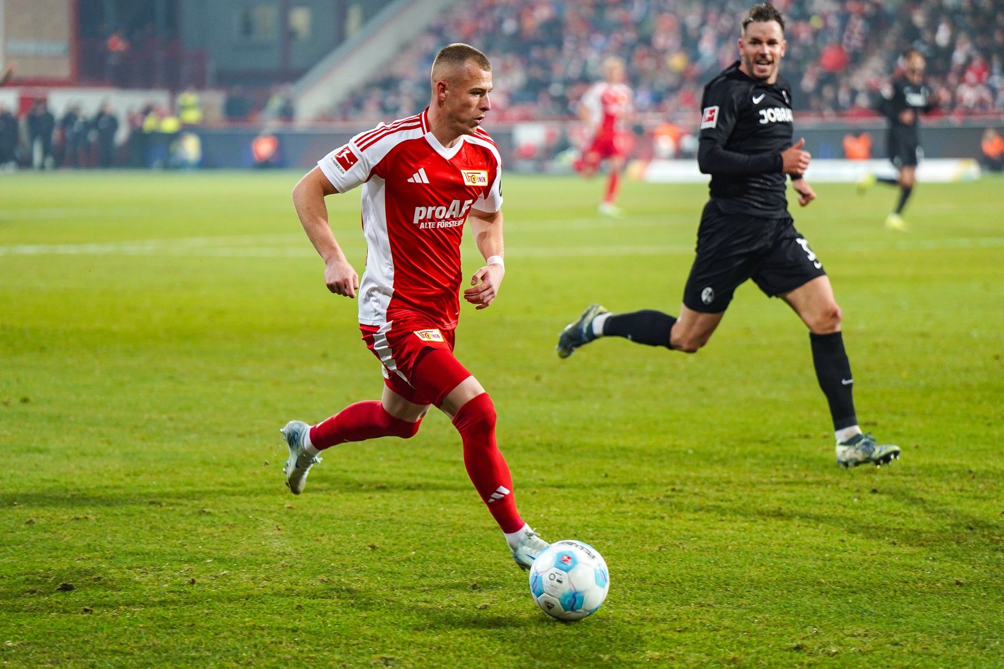 A football player in a red uniform is dribbling the ball on a stadium field while an opposing player in black is chasing him.