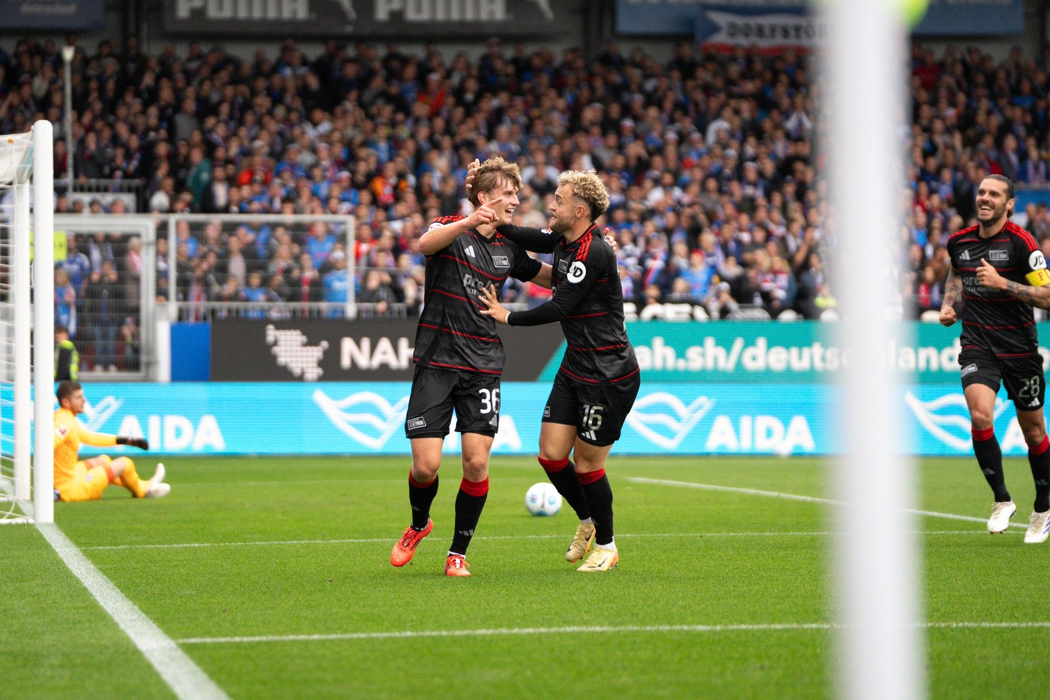 Zwei Fußballspieler umarmen sich freudig nach einem Tor in einem Stadion mit jubelnden Fans im Hintergrund.