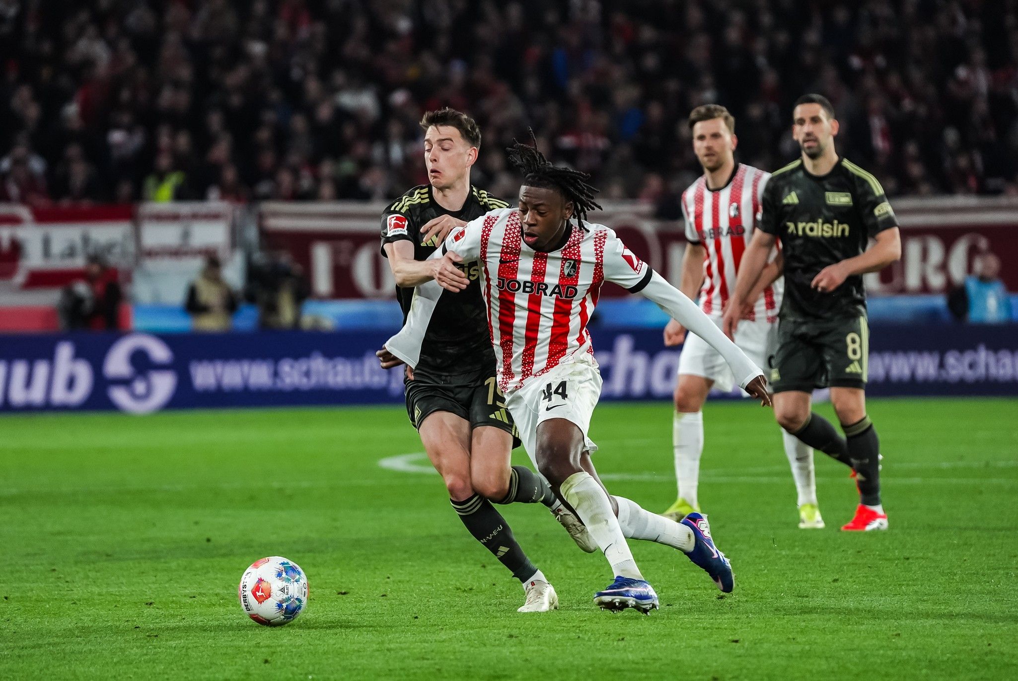 Ein Fußballspieler mit der Nummer 14 in rot-weiß gestreifter Kleidung dribbelt den Ball, während ein Gegner ihn verfolgt.