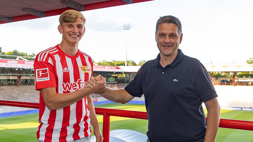 Young soccer player in a striped jersey shakes hands with a man in front of a stadium with stands in the background.