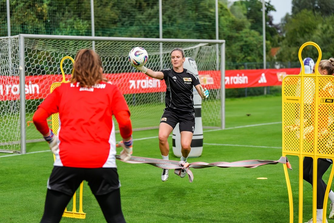 Eine Fußballspielerin springt über ein Hindernis, während eine andere Spielerin im Hintergrund den Ball fängt.
