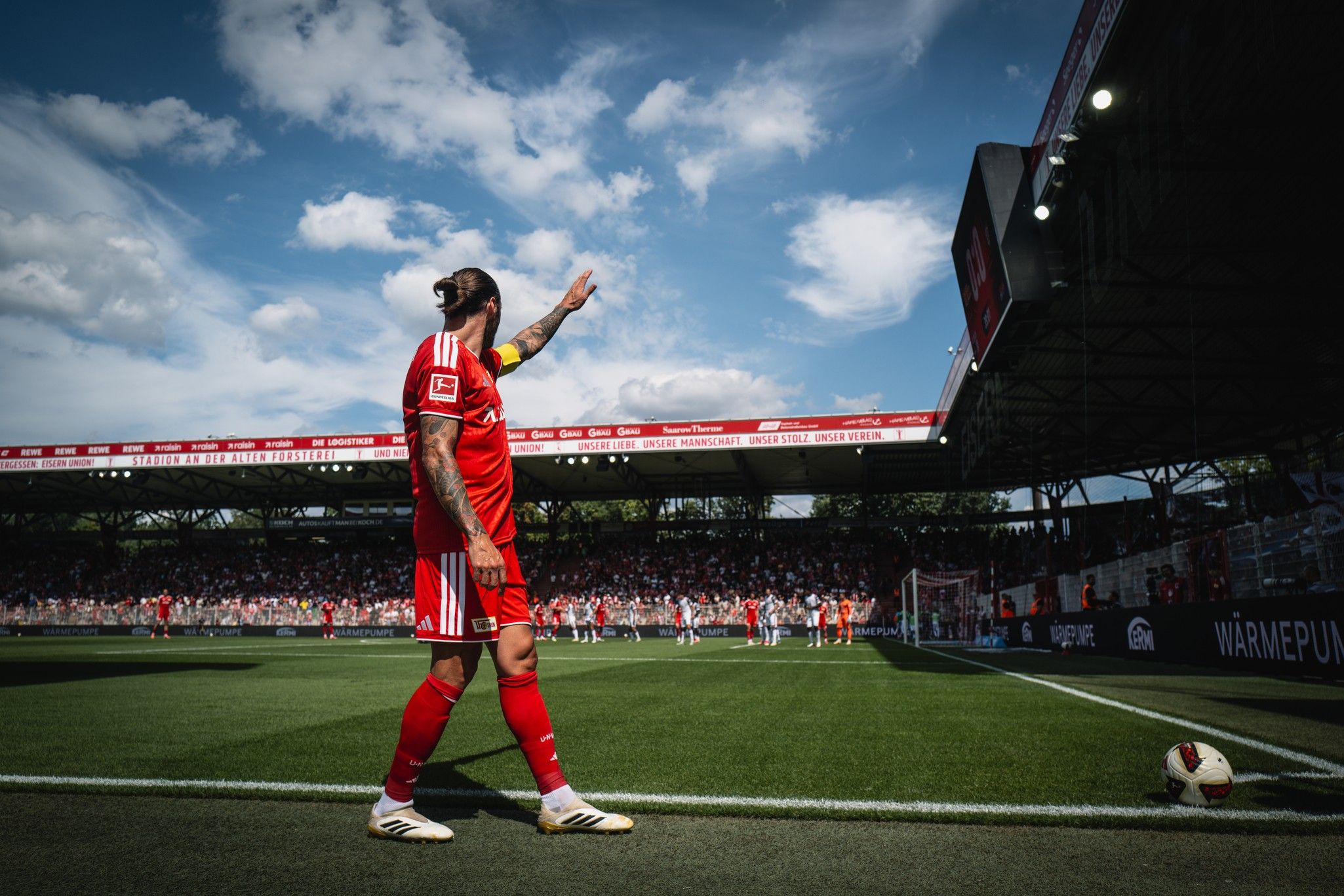 Ein Fußballspieler in rotem Trikot steht an der Seitenlinie und hebt die Hand, während im Hintergrund ein Stadion voller Zuschauer zu sehen ist.