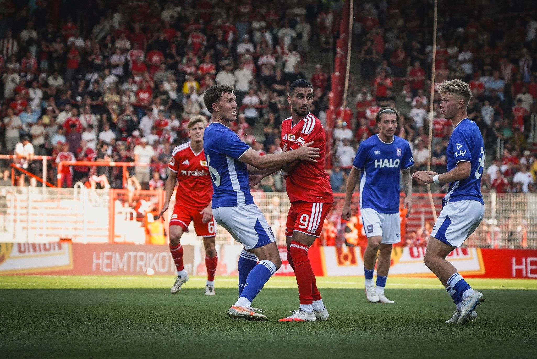 Zwei Fußballspieler in roten und blauen Trikots kämpfen um den Ball, umgeben von jubelnden Zuschauern im Stadion.