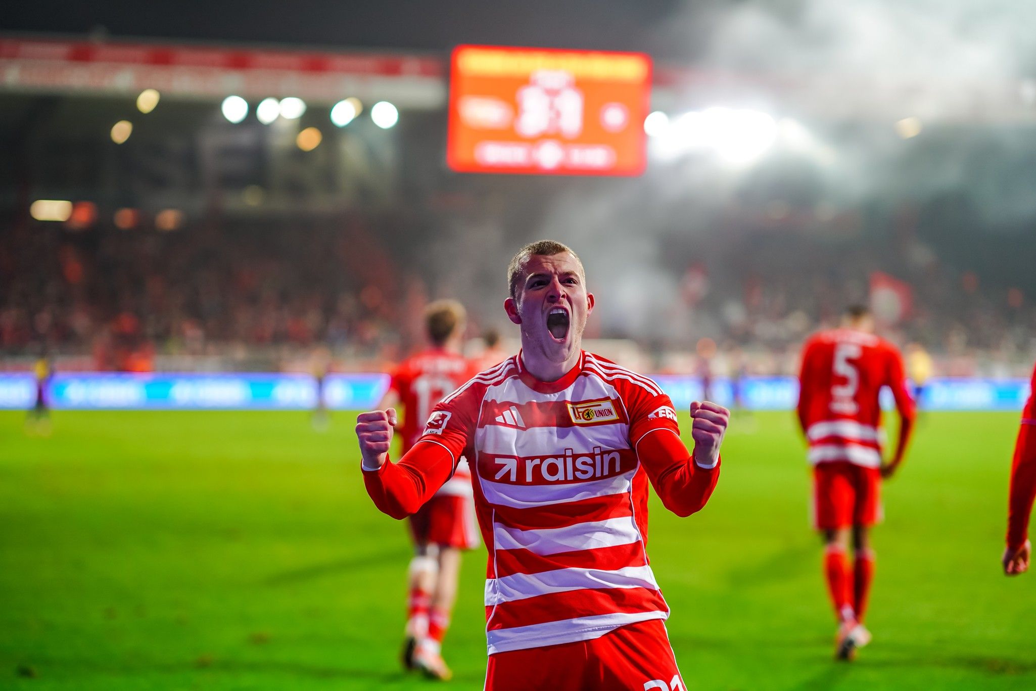 Spieler in rot-weißer Fußballausrüstung jubelt auf dem Feld, im Hintergrund jubelnde Fans und Stadionbeleuchtung.