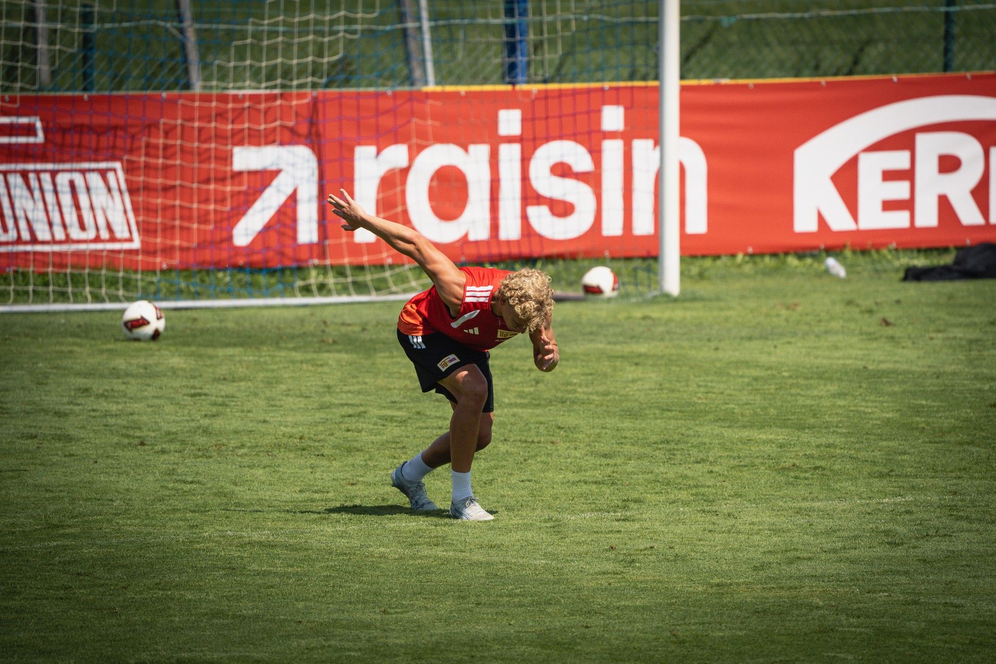 Ein Spieler in rotem Trikot führt eine Bewegungsübung auf einem Fußballfeld durch, im Hintergrund Netz und Bandenwerbung.
