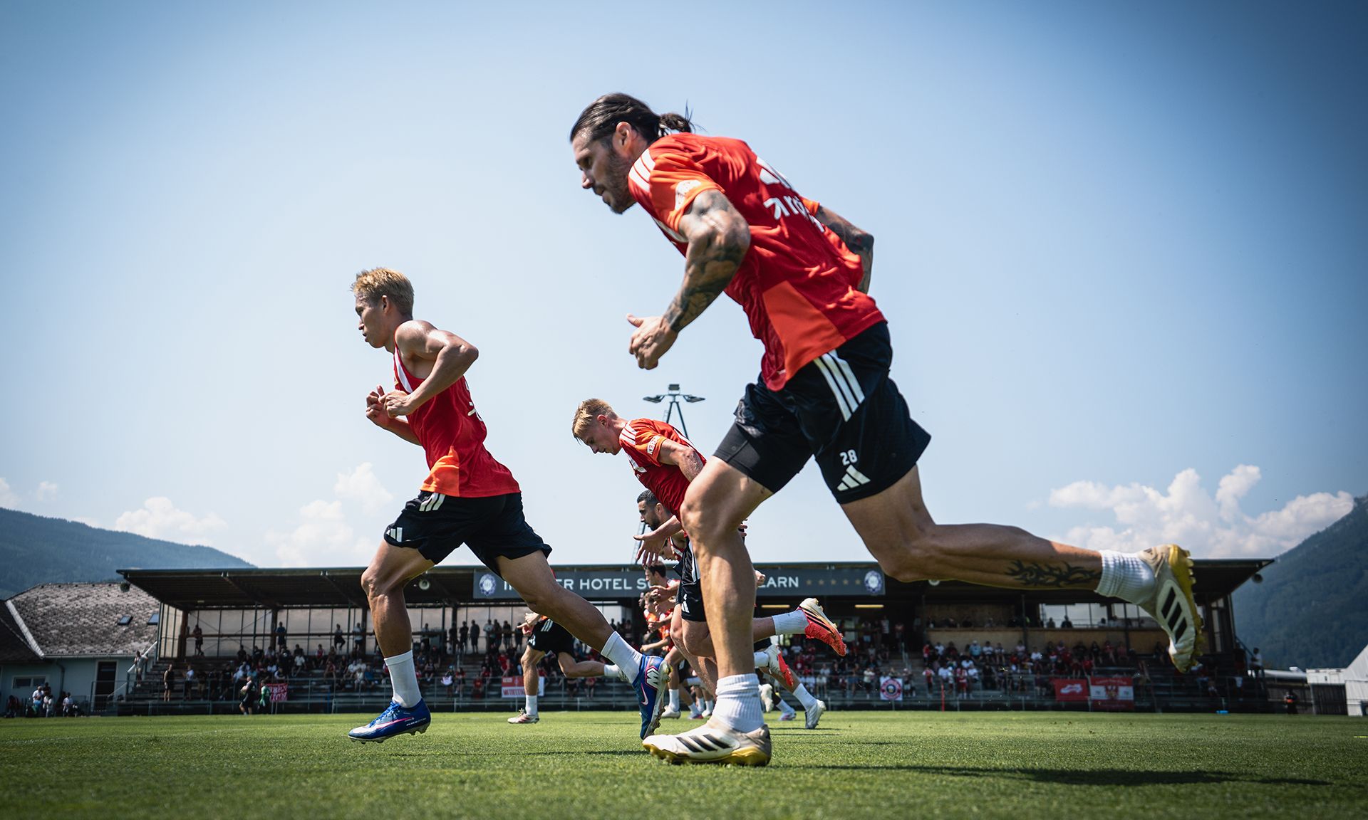 Spieler in roten Trikots trainieren auf einem Sportplatz bei sonnigem Wetter, während Zuschauer im Hintergrund zuschauen.