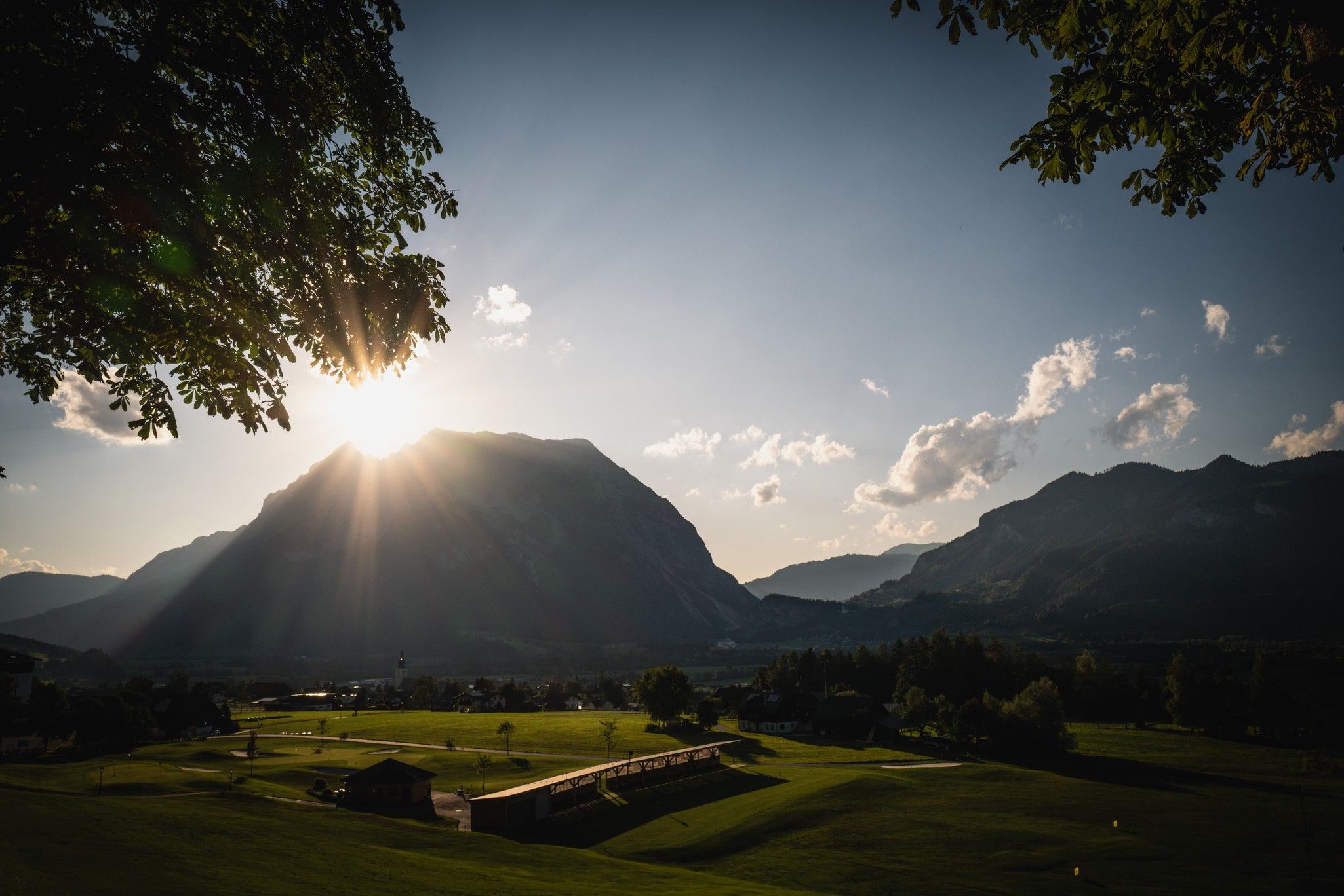 Sonnenuntergang hinter einem Berg, der Lichtstrahlen wirft, umgeben von grünen Wiesen und bewaldeten Hügeln.