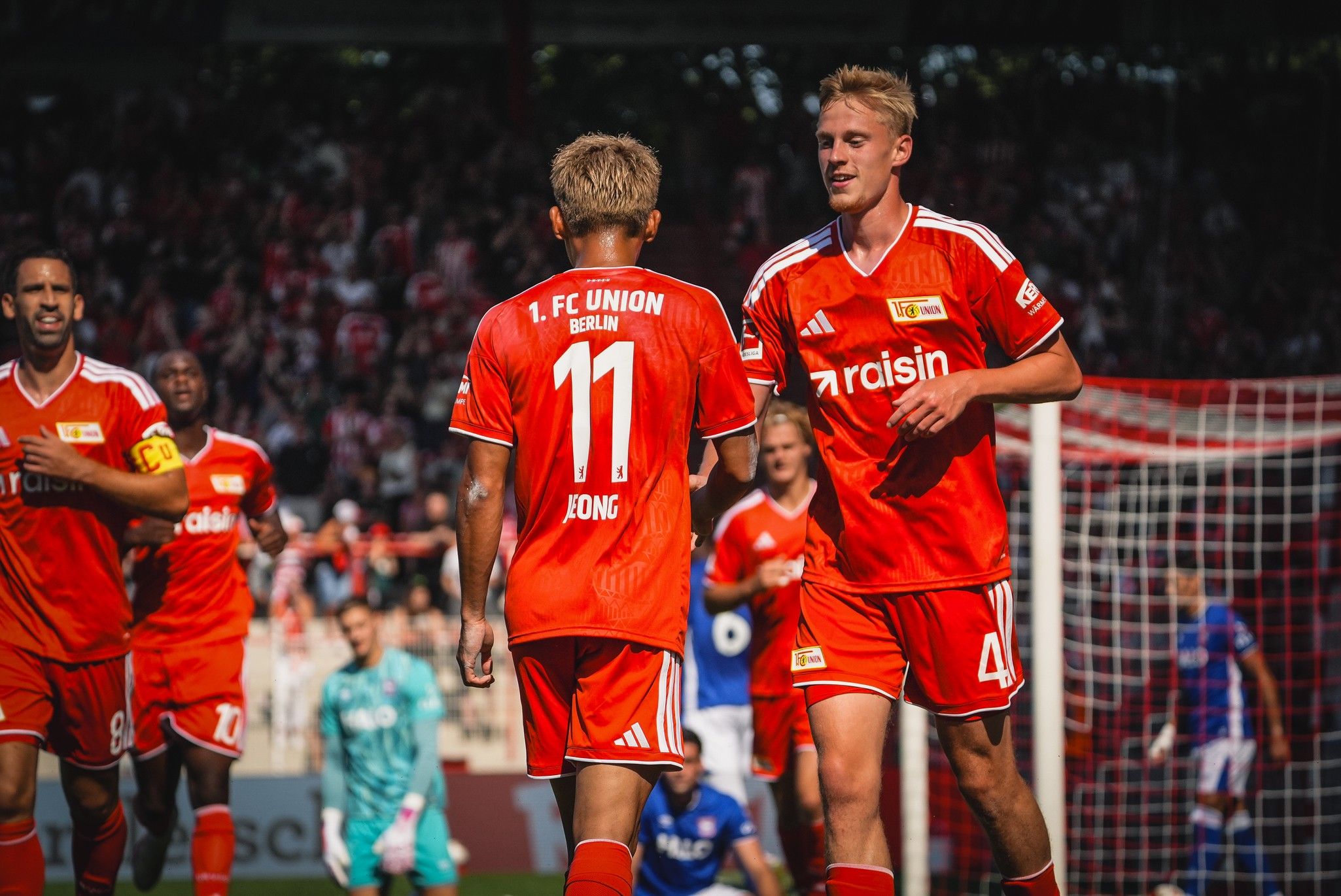 Zwei Spieler des 1. FC Union Berlin in roten Trikots feiern ein Tor auf dem Spielfeld, mit Zuschauern im Hintergrund.
