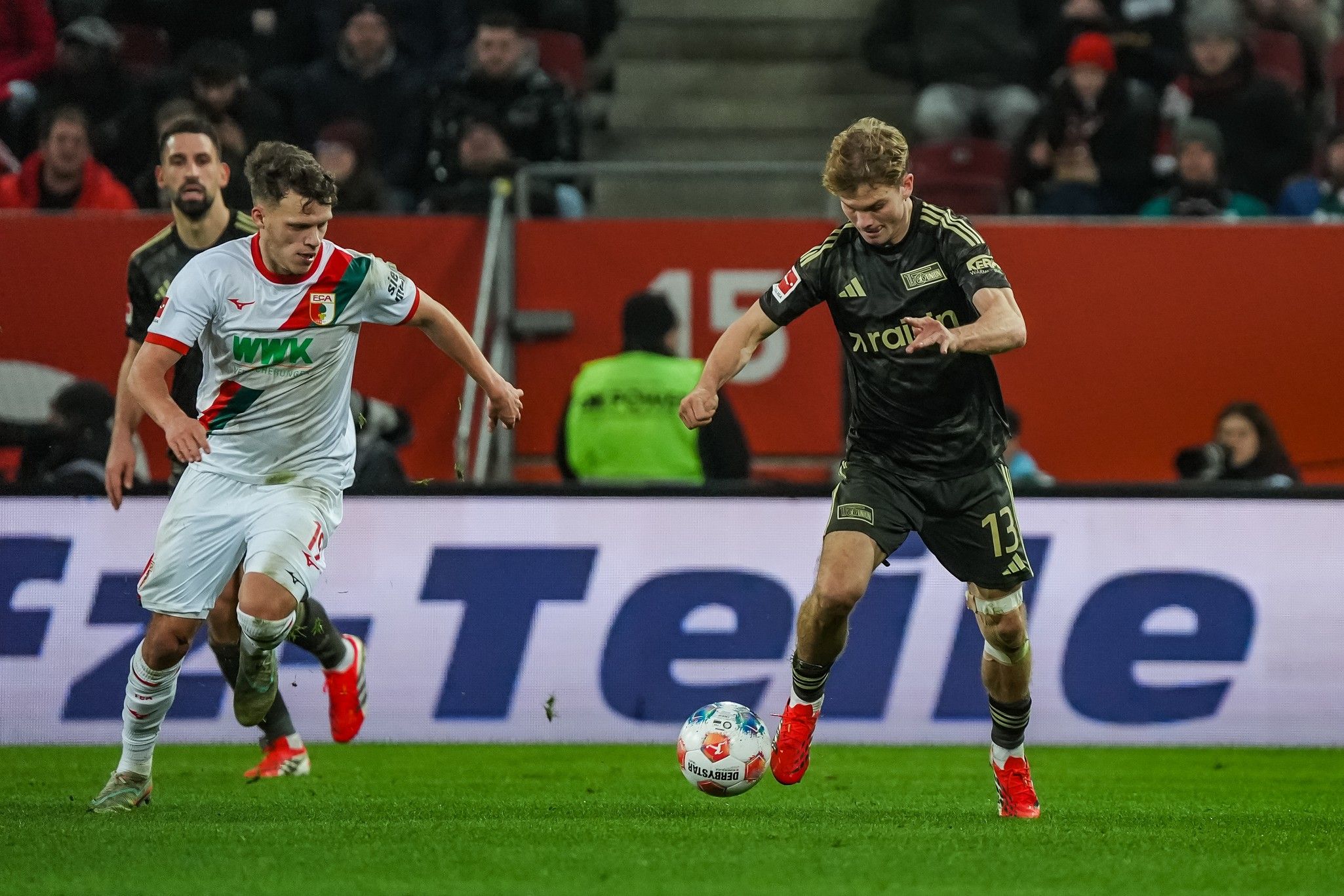 Zwei Fußballspieler im Wettkampf um den Ball während eines Spiels in einem Stadion, Zuschauer im Hintergrund.