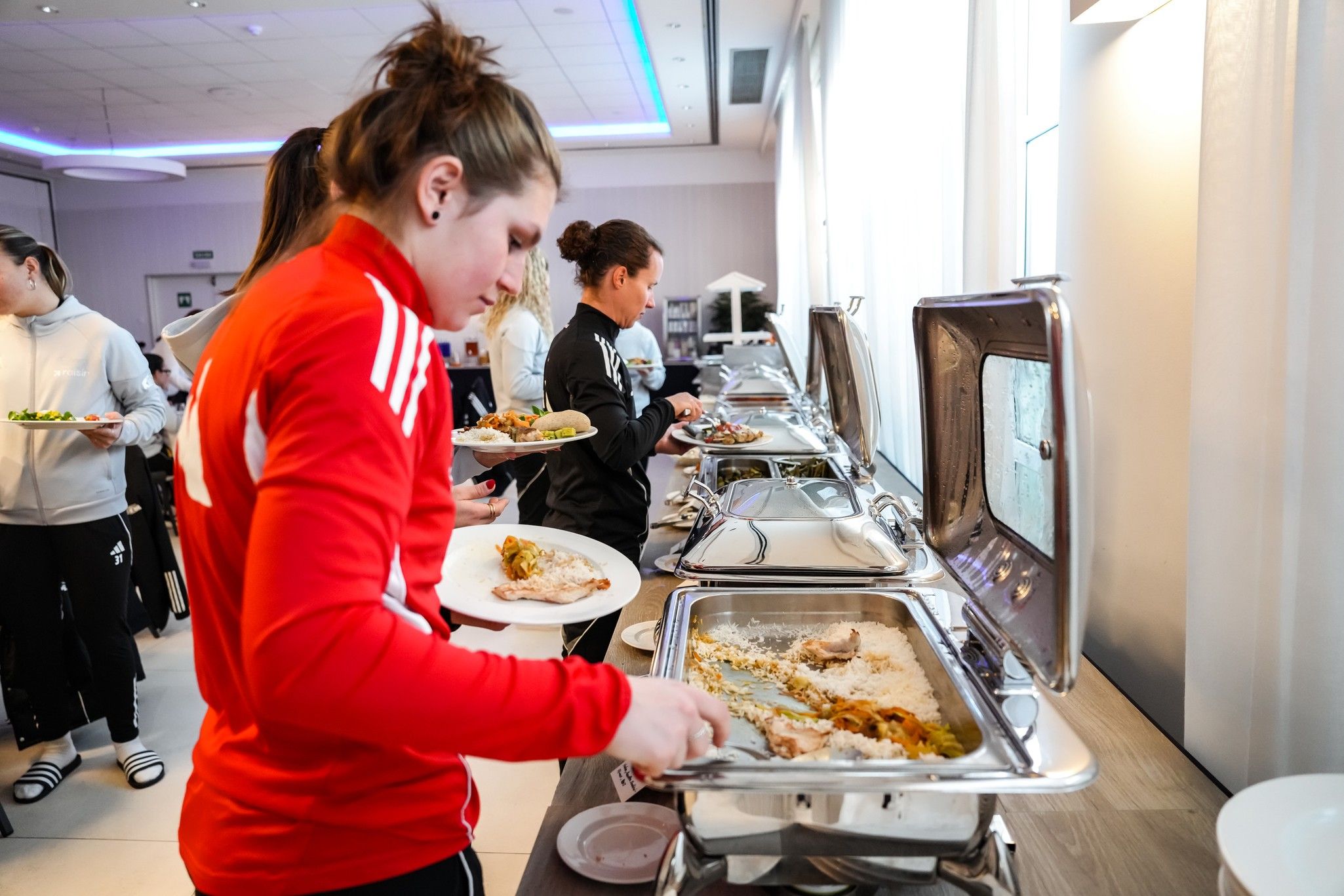 Eine Person in roter Sportjacke befüllt ihren Teller an einem Buffet mit Essen, während andere im Hintergrund ebenfalls Speisen auswählen.