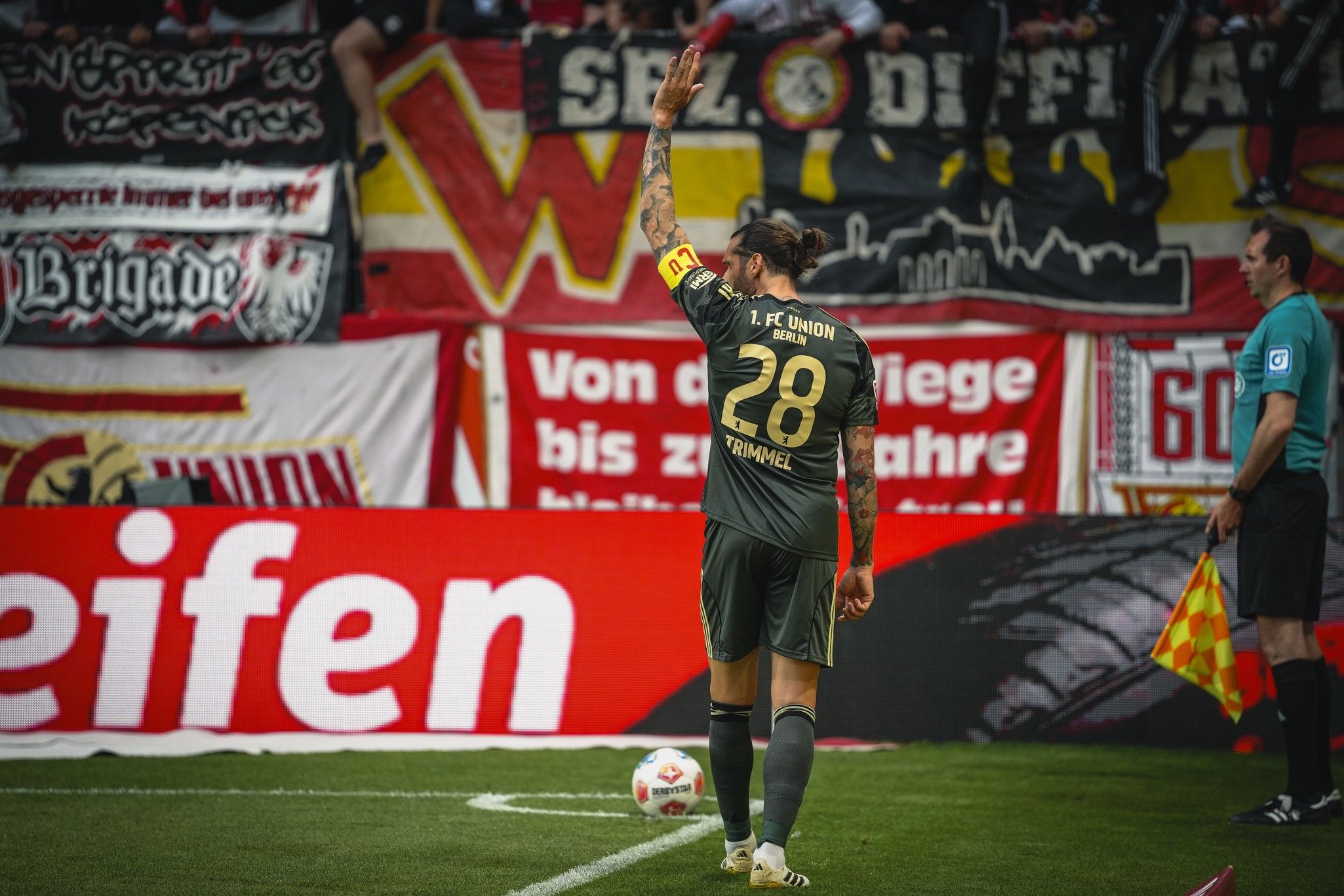 Ein Fußballspieler mit der Nummer 28 winkt von der Seitenlinie, im Hintergrund sind Fanbanner und ein Schiedsrichter zu sehen.