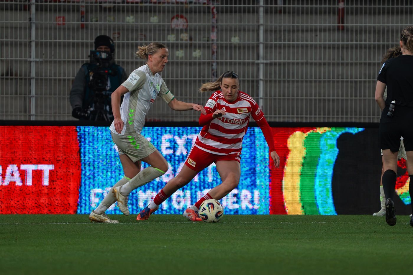 Zwei Frauen im Fußballspiel: Eine Spielerin in rot-weiß gestreiftem Trikot dribbelt, während die Gegenspielerin sie verfolgt.