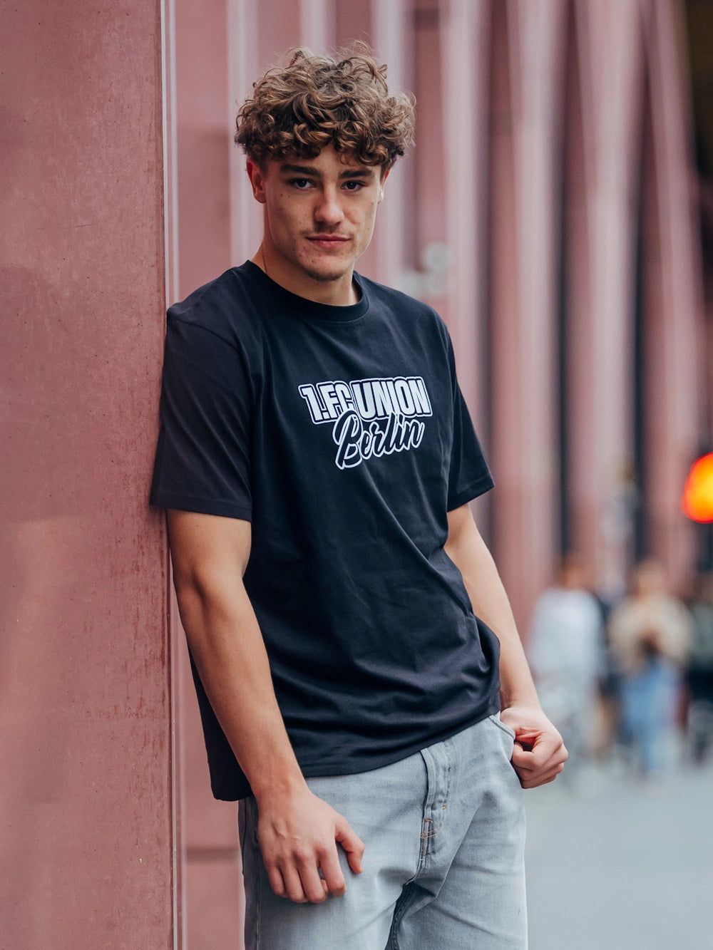 Junger Mann mit lockigem Haar, schwarzem T-Shirt mit Schriftzug und Jeans steht an einer Wand in einer urbanen Umgebung.